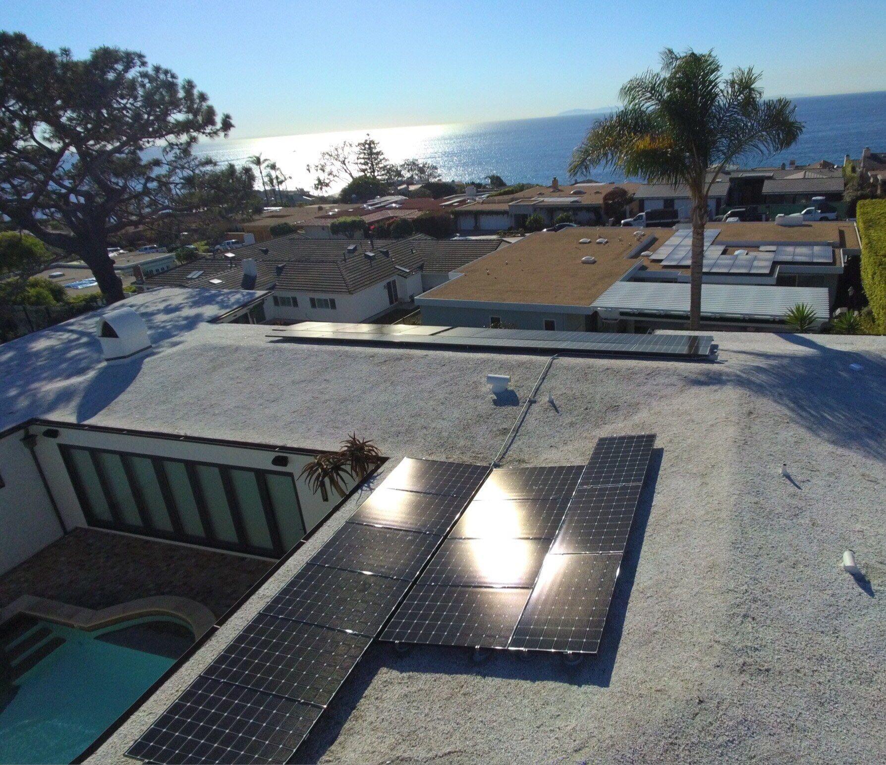 Solar panels glinting in afternoon sun on a coastal flat roof, with a palm tree and sunlight reflecting off the Pacific Ocean behind