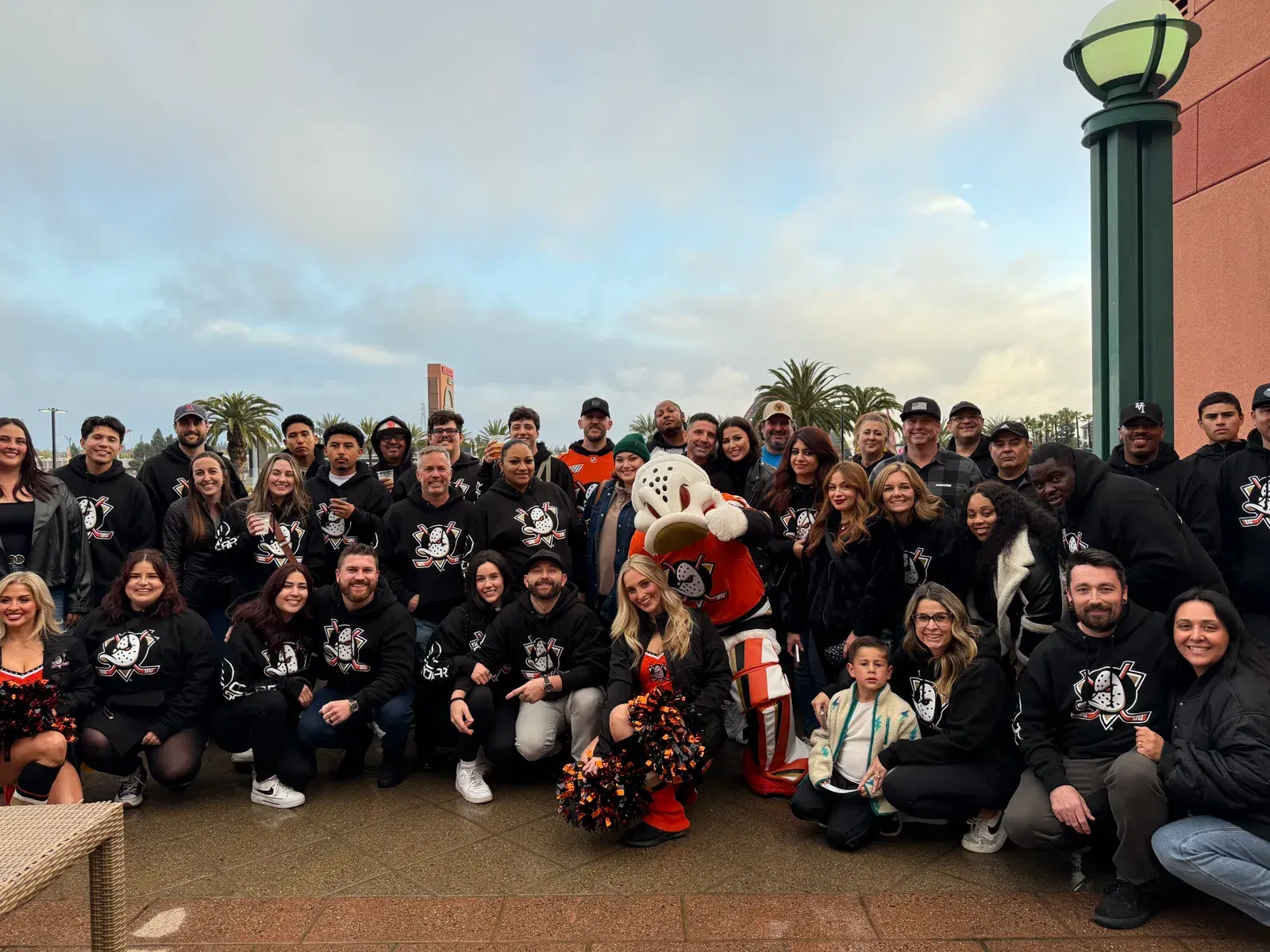A large group of OC Solar employees in matching Anaheim Ducks sweatshirts pose outdoors with the Ducks mascot Wild Wing in a goalie uniform at Honda Center.
