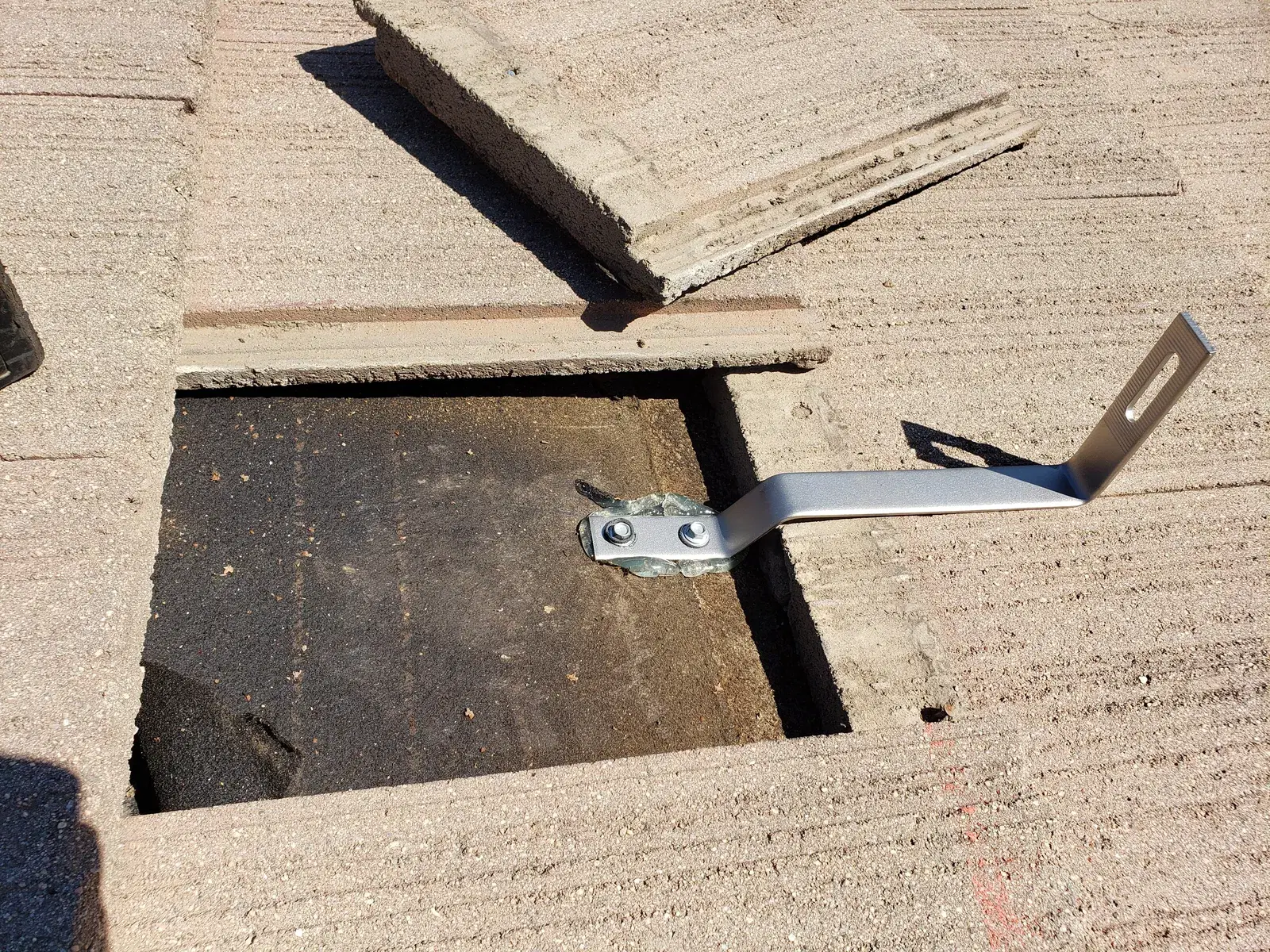 Close-up of a metal solar panel mounting bracket bolted to a roof deck with concrete tiles pulled back to expose the underlayment.