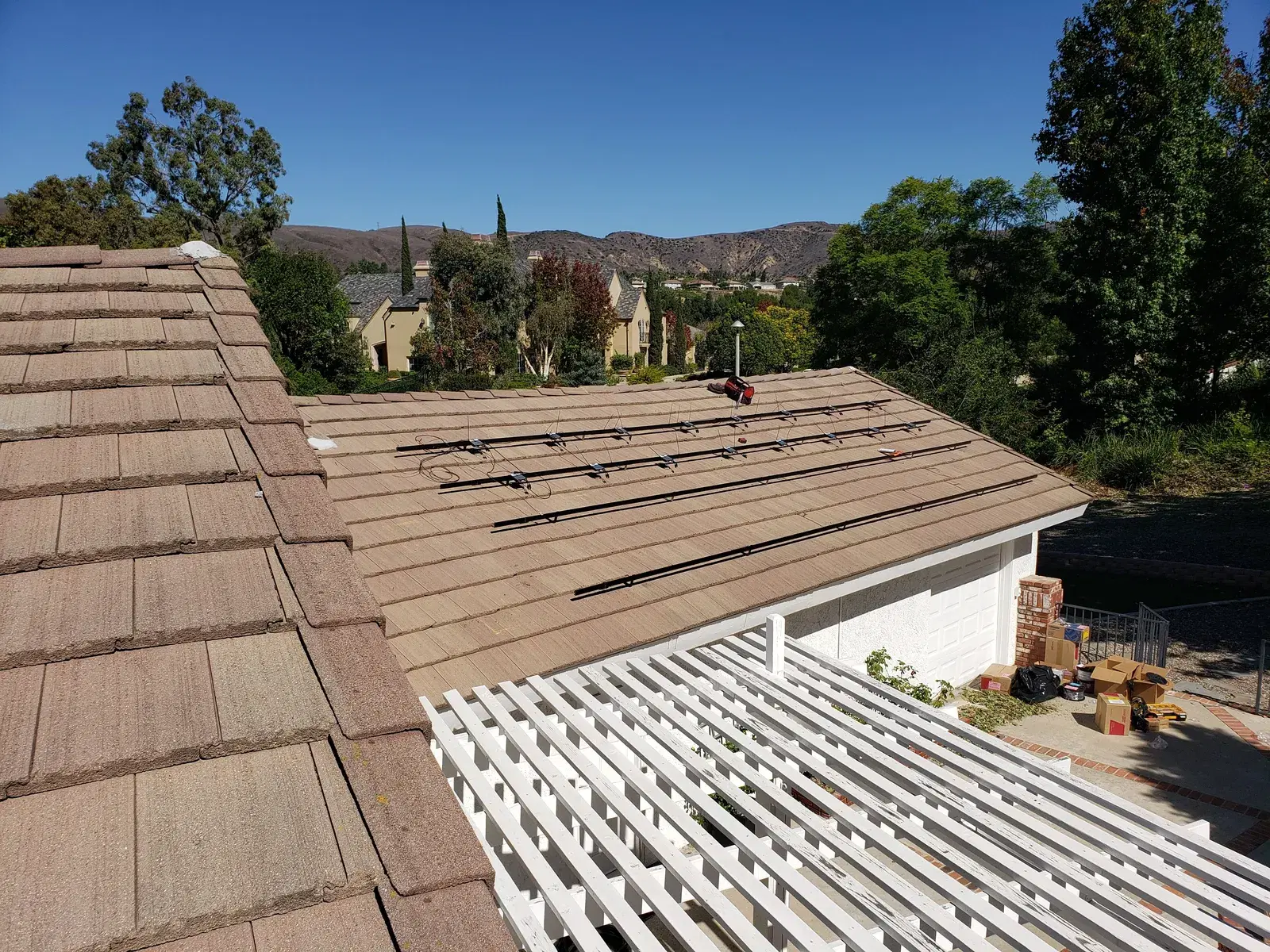 A residential tile roof with solar mounting rails and black wiring runs installed, set against blue sky and distant green hills.