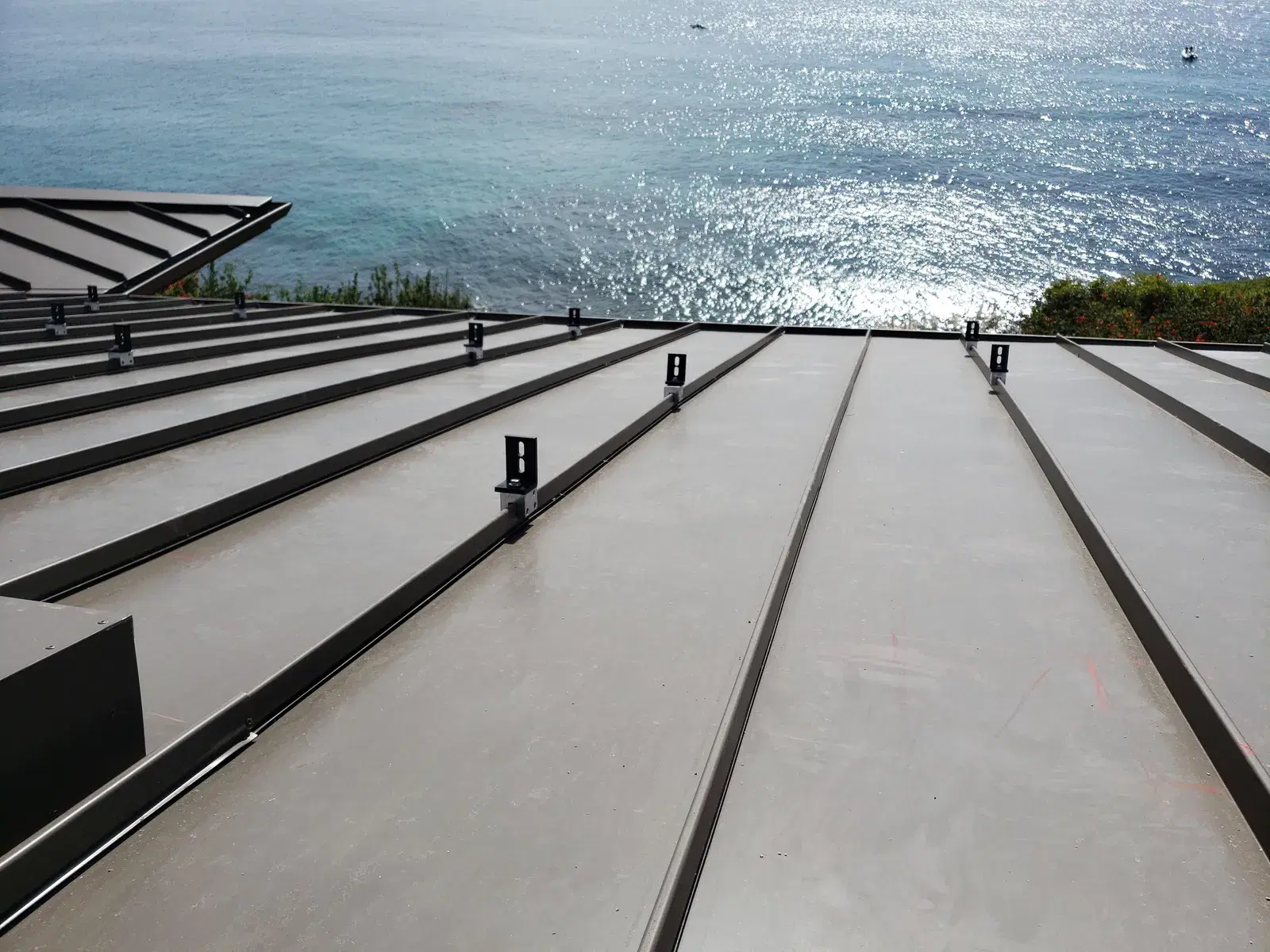 A standing-seam metal roof fitted with black solar mounting brackets, overlooking a sunlit ocean.