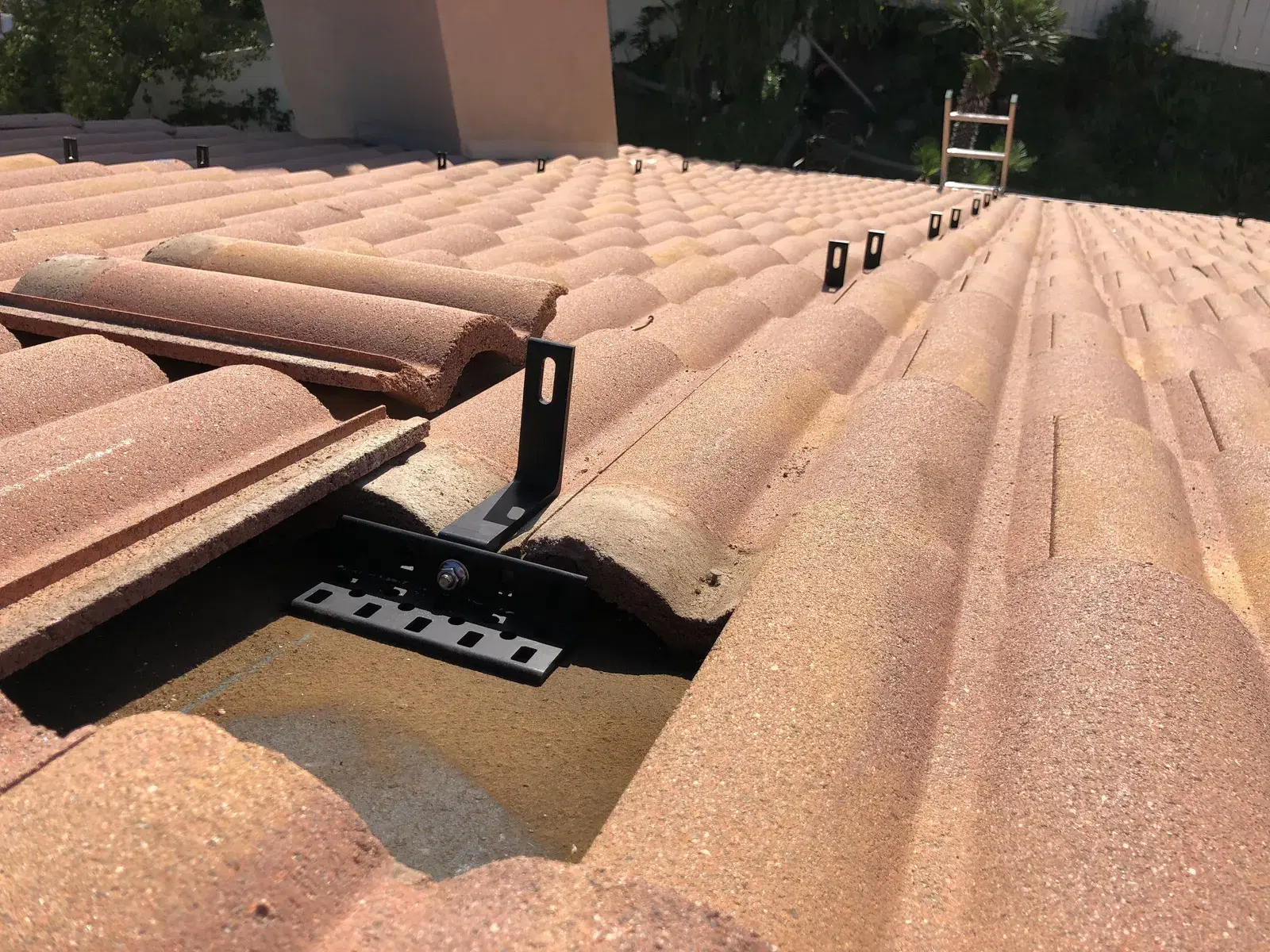 Close-up of a black metal solar panel mounting foot and rail bracket installed under a raised barrel clay roof tile, with more mounting hooks visible along the tile roof.