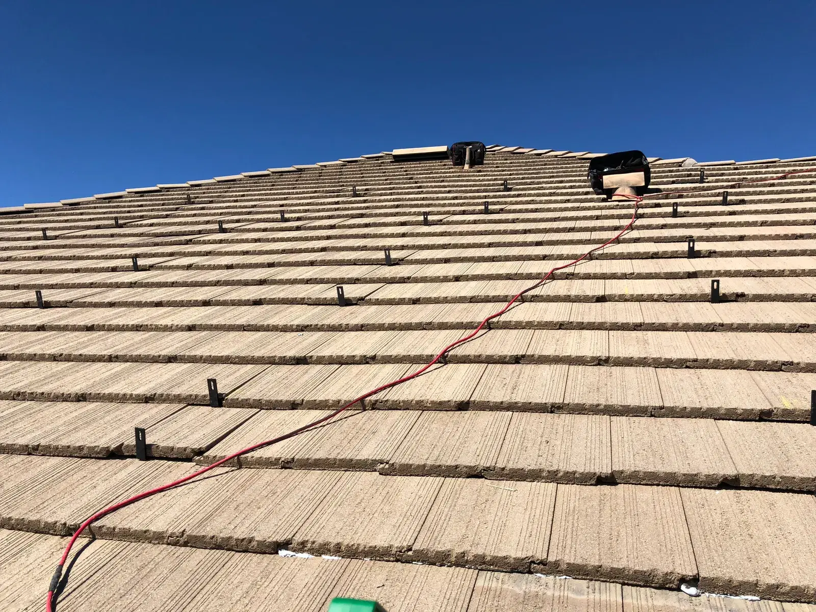 Tile roof with rows of black solar mounting feet installed and a red electrical wire running diagonally up toward the roof ridge against a bright blue sky.
