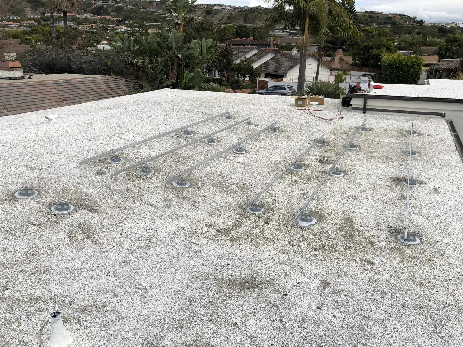 A flat white rooftop with rows of metal solar mounting feet and rails arranged in a diagonal grid, with houses and palm trees in the background.