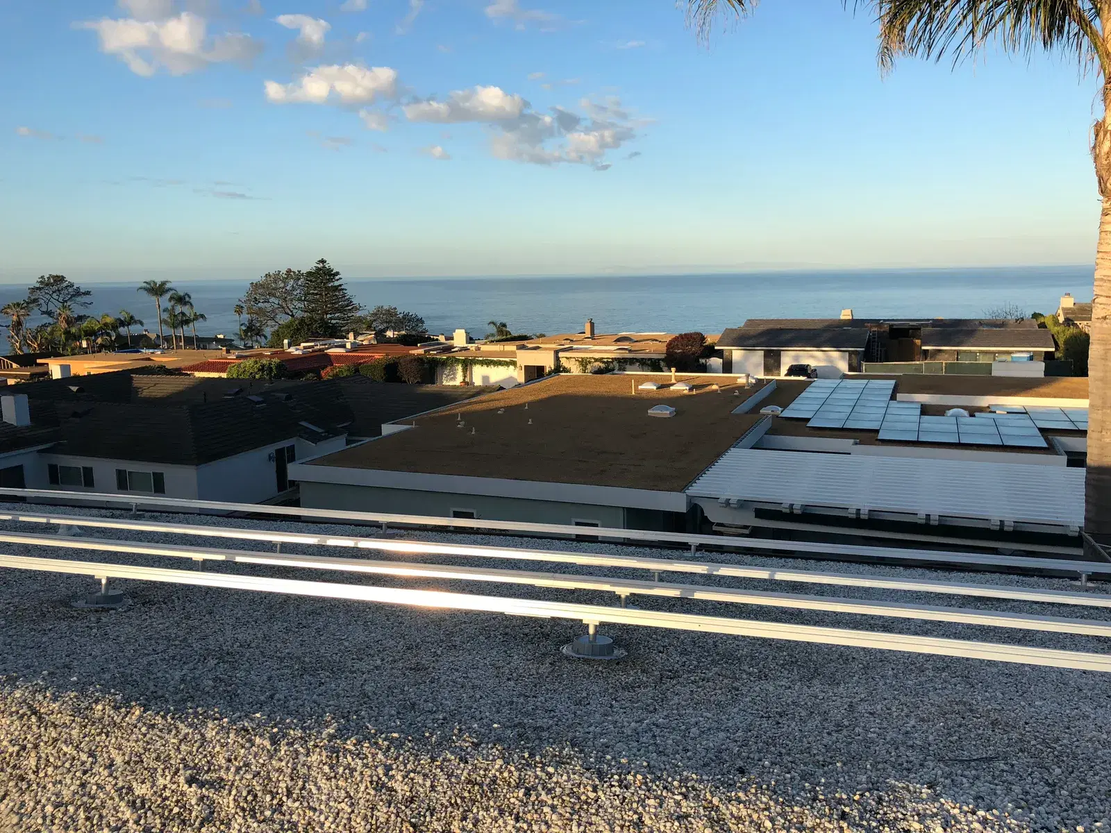 A flat rooftop with solar mounting rails in the foreground and a solar array on a nearby roof, with ocean-view homes and the sea beyond.