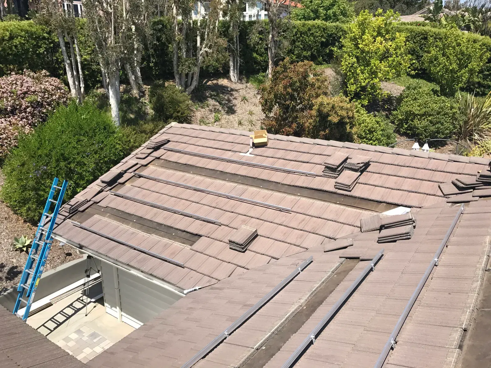 A concrete-tile roof with solar mounting rails laid out and tiles lifted, a blue ladder propped against the eave.