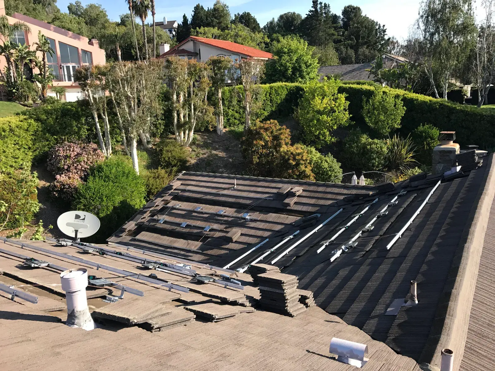 A concrete-tile roof with rows of solar mounting rails and standoffs installed, garden and homes visible behind.
