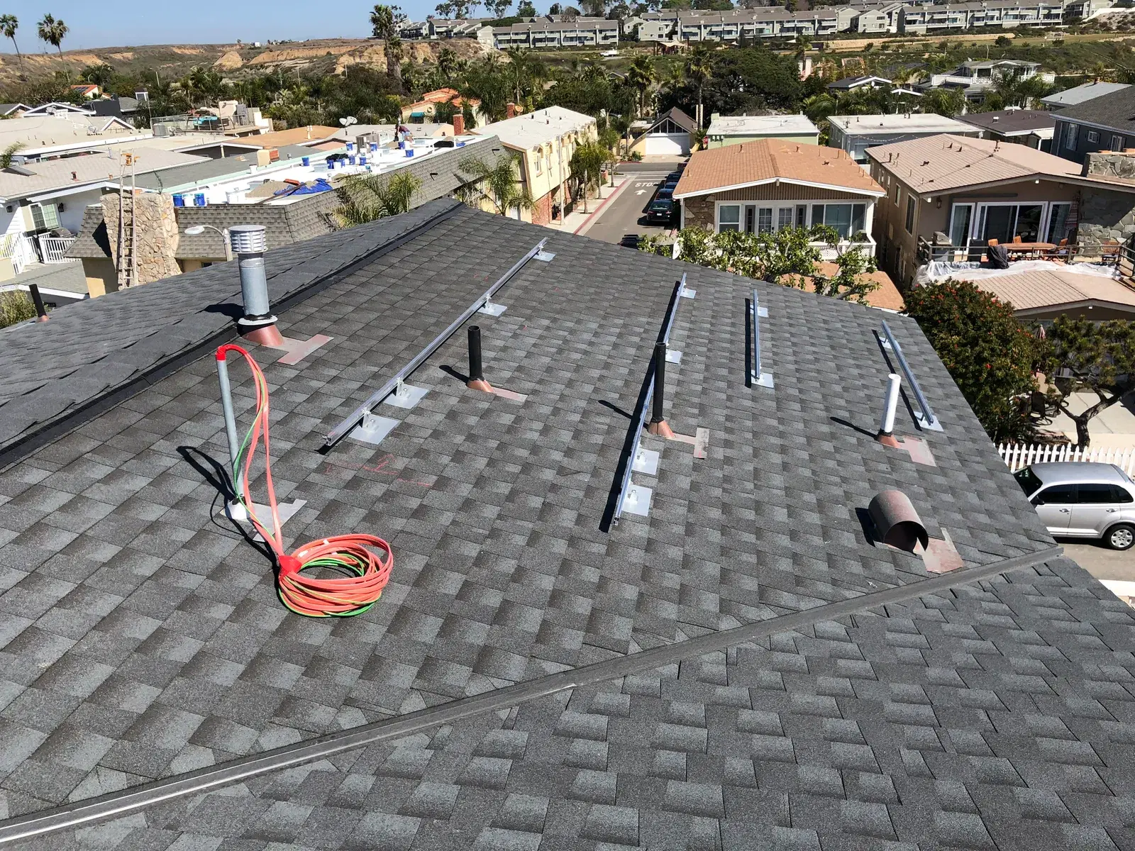 An asphalt-shingle residential rooftop with solar mounting rails and standoffs laid out and an orange extension cord coiled nearby, overlooking a hillside neighborhood.