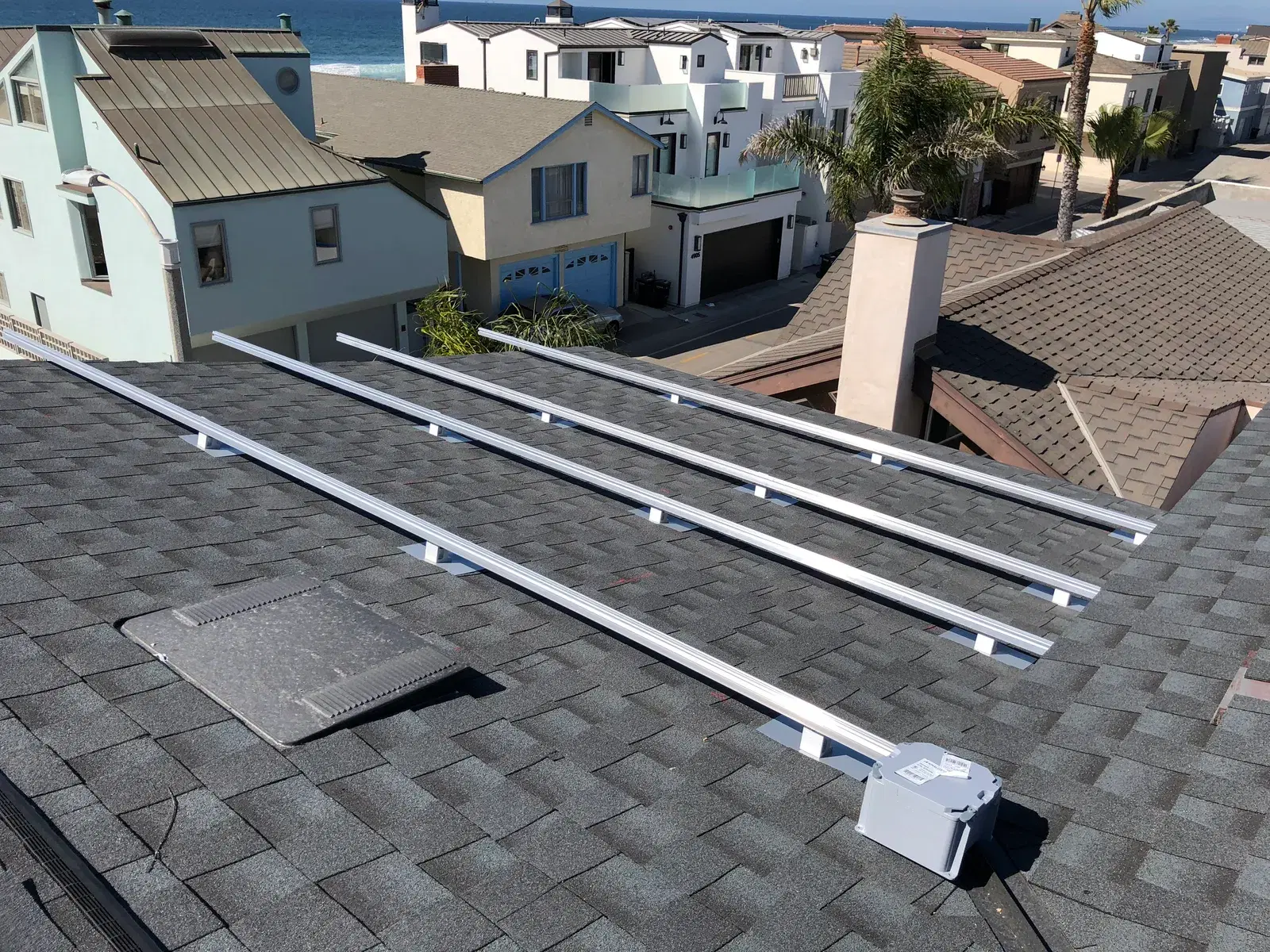 Rows of newly installed aluminum mounting rails and a white junction box on a gray composition-shingle roof, with beach houses and the ocean visible in the background.