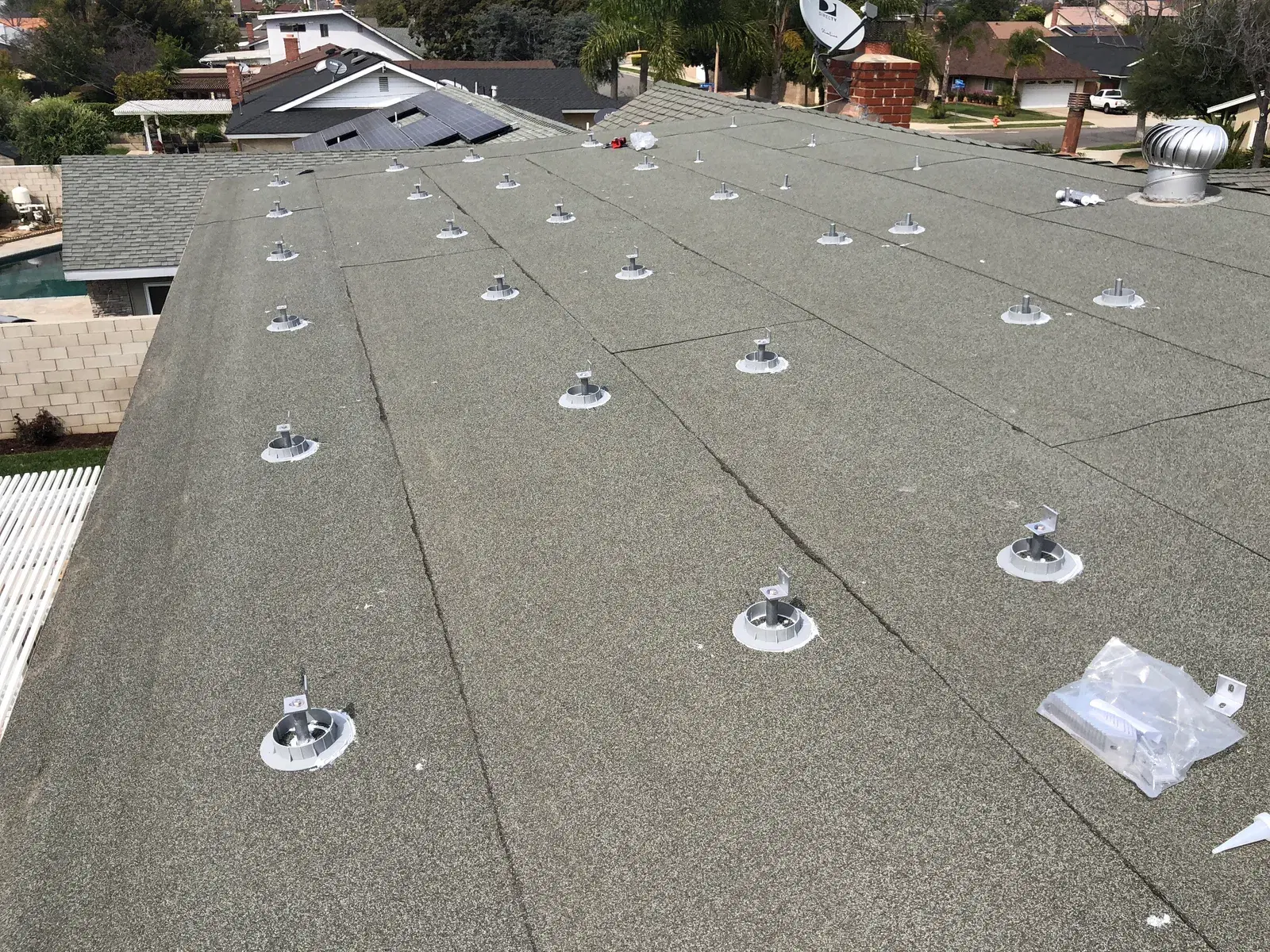 A residential shingle rooftop with multiple metal mounting standoffs and flashings installed in rows, plus a bag of mounting hardware, in preparation for solar panel racking.
