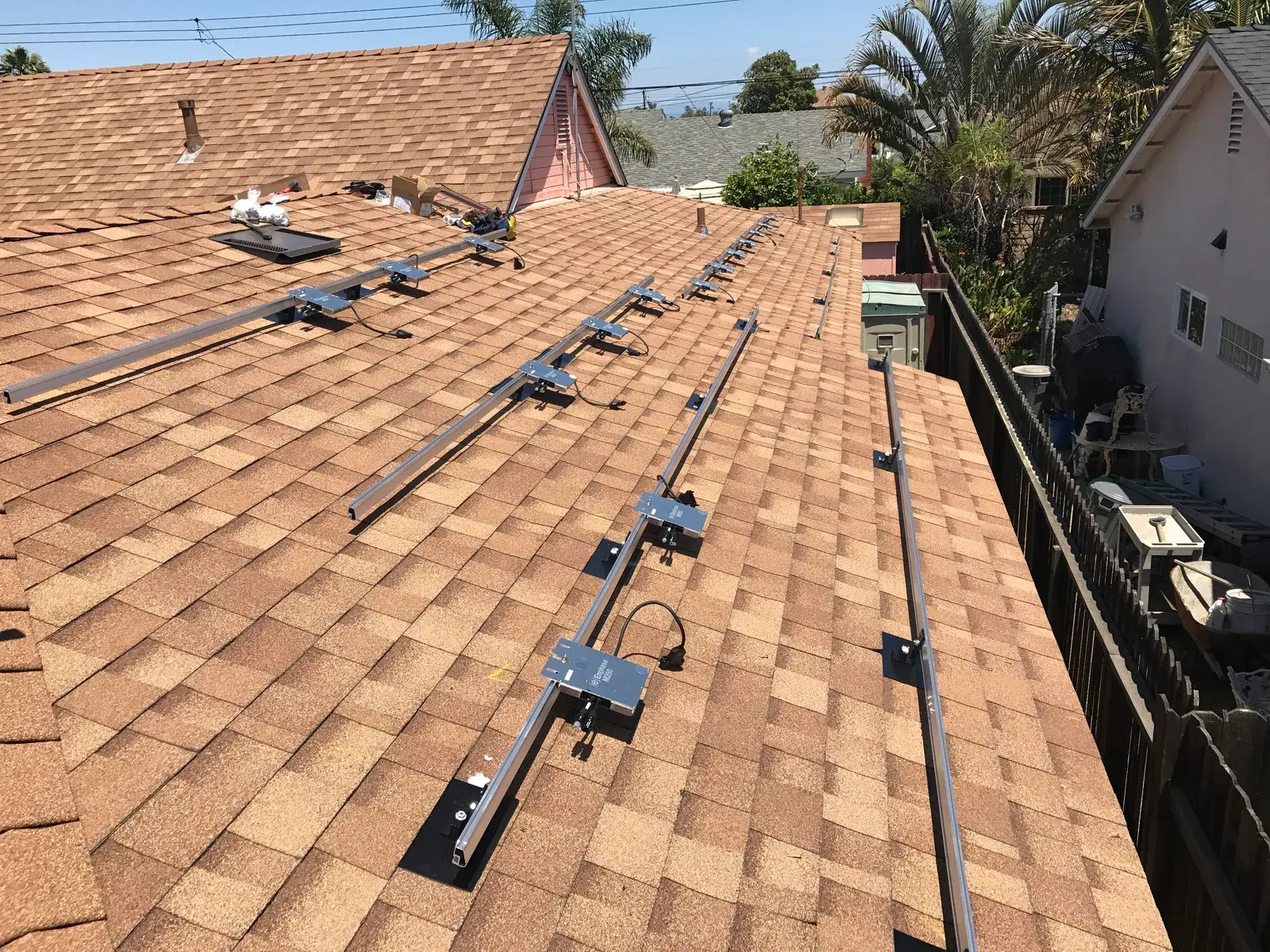 A residential shingled rooftop with aluminum solar mounting rails and attachment feet installed in rows, awaiting solar panel placement, with neighborhood homes and palm trees in the background.