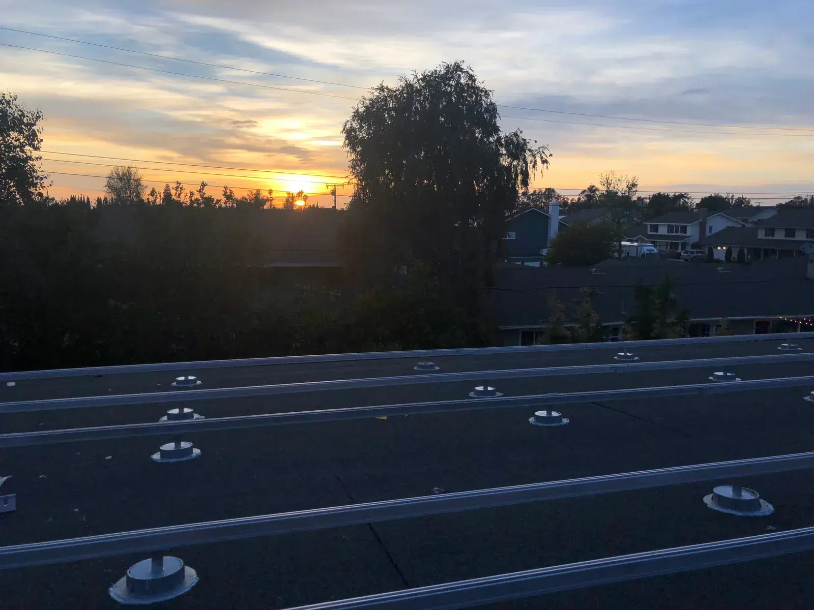 Rows of metal solar mounting rails and standoff feet on a flat rooftop at dusk, with neighborhood houses and an orange sunset in the background.