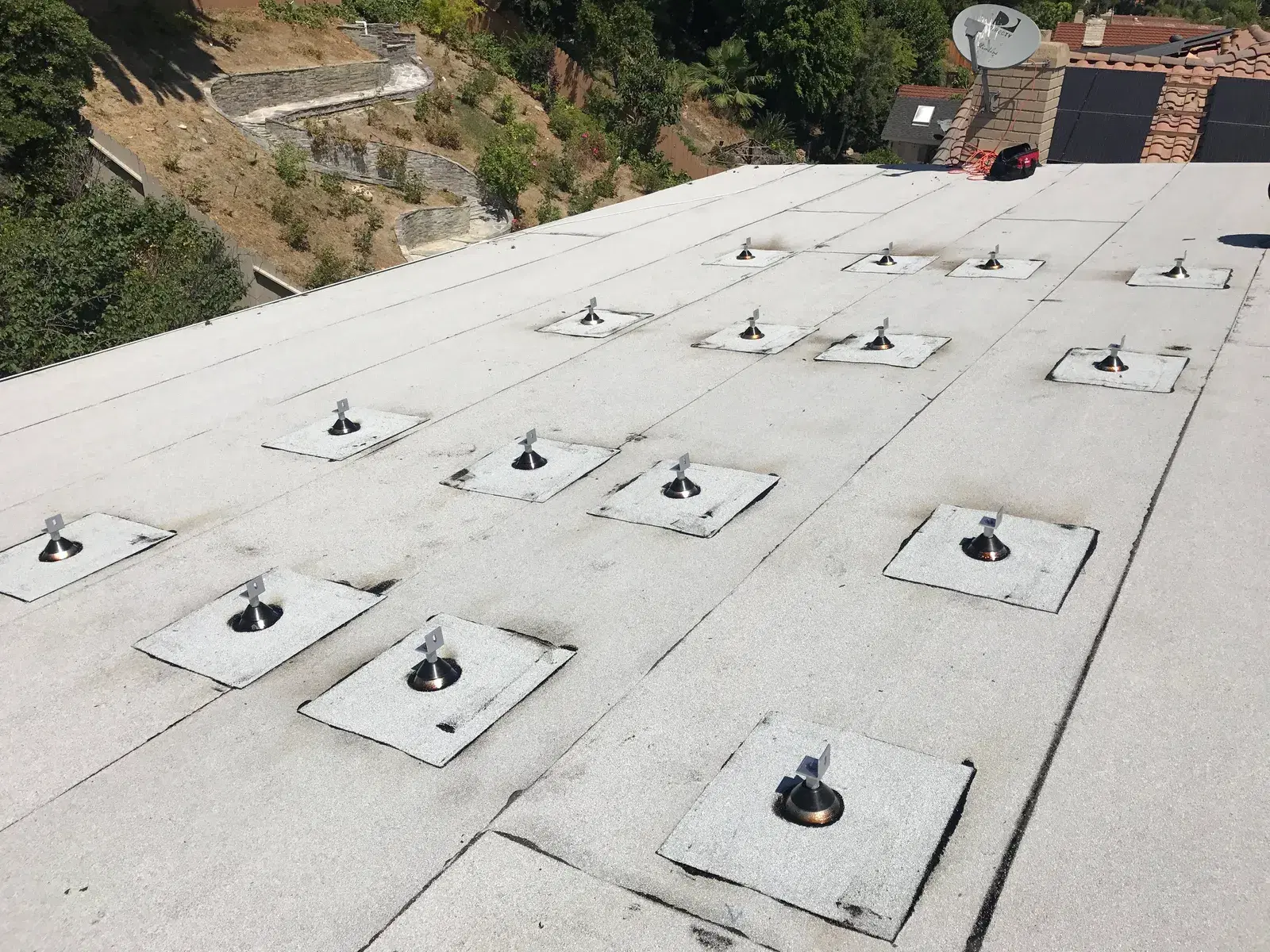A flat residential rooftop with a grid of metal solar mounting feet and flashings installed, awaiting panels, overlooking a green hillside.