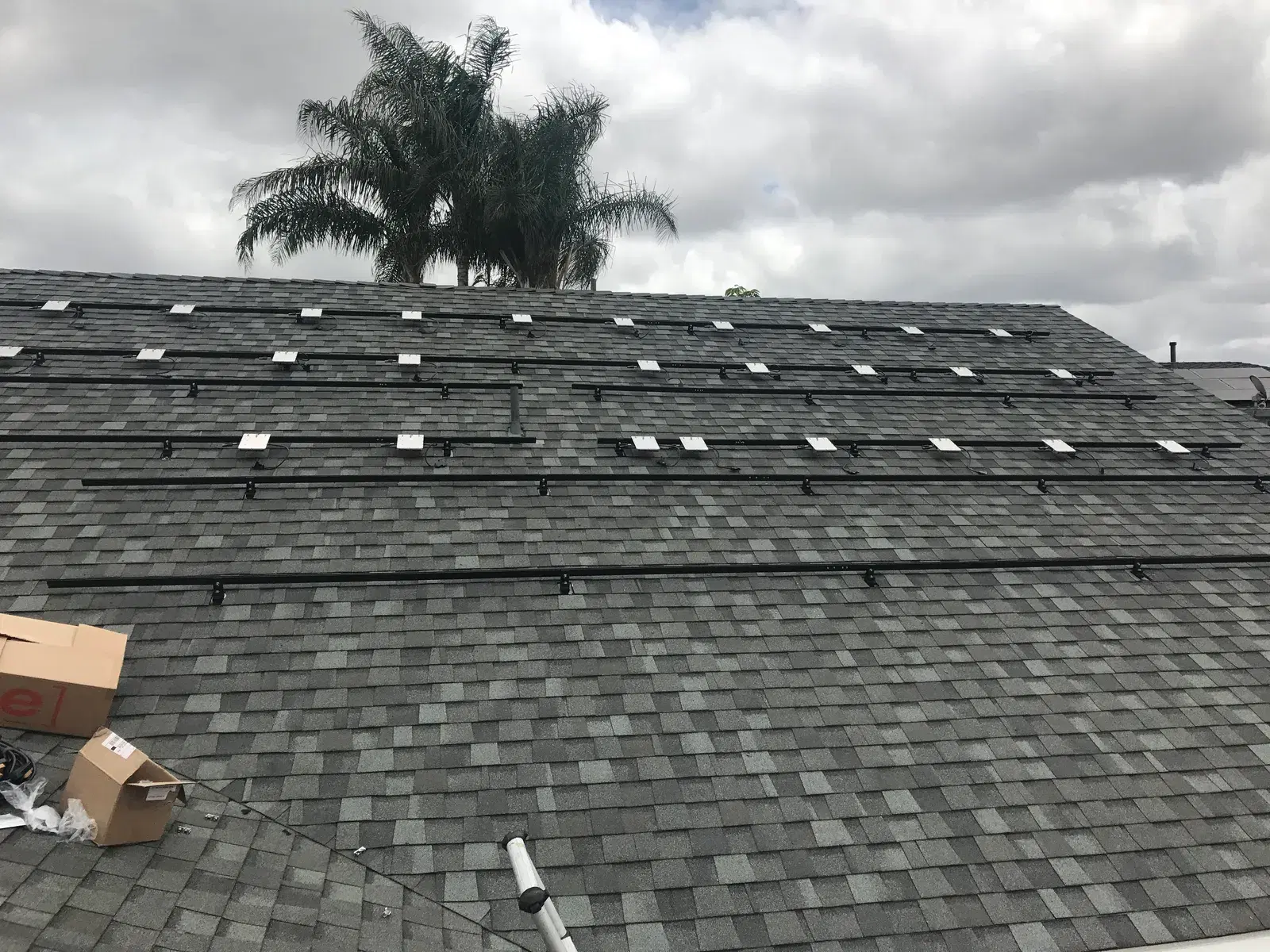 A residential gray shingle roof fitted with rows of black solar mounting rails and attachment feet, with a palm tree and cloudy sky in the background.