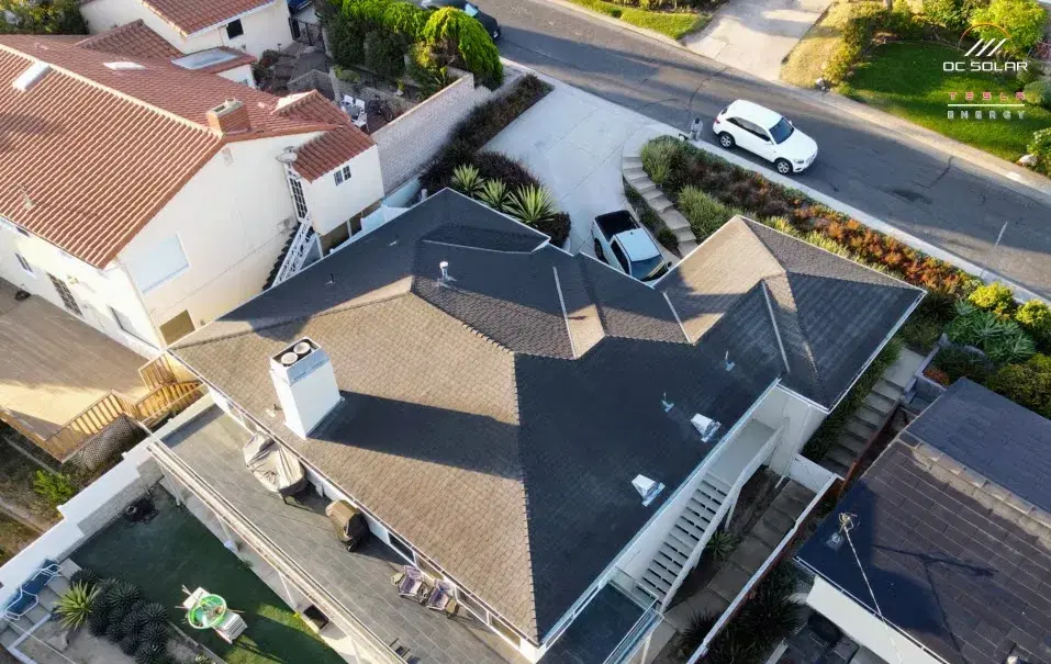 Drone view of a multi-gable shingle roof with mounting hardware staged across it, surrounded by a neighborhood street and yards.