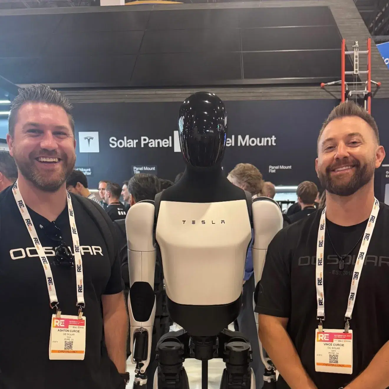Two smiling men in OC Solar shirts with conference lanyards stand on either side of a black-and-white Tesla Optimus robot at a trade show booth.