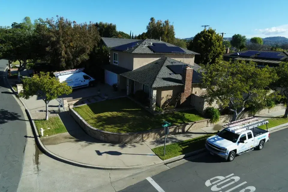 Aerial view of a two-story suburban house with a branded solar company van in the driveway and a branded pickup truck parked at the corner curb, with rooftop solar panels visible.
