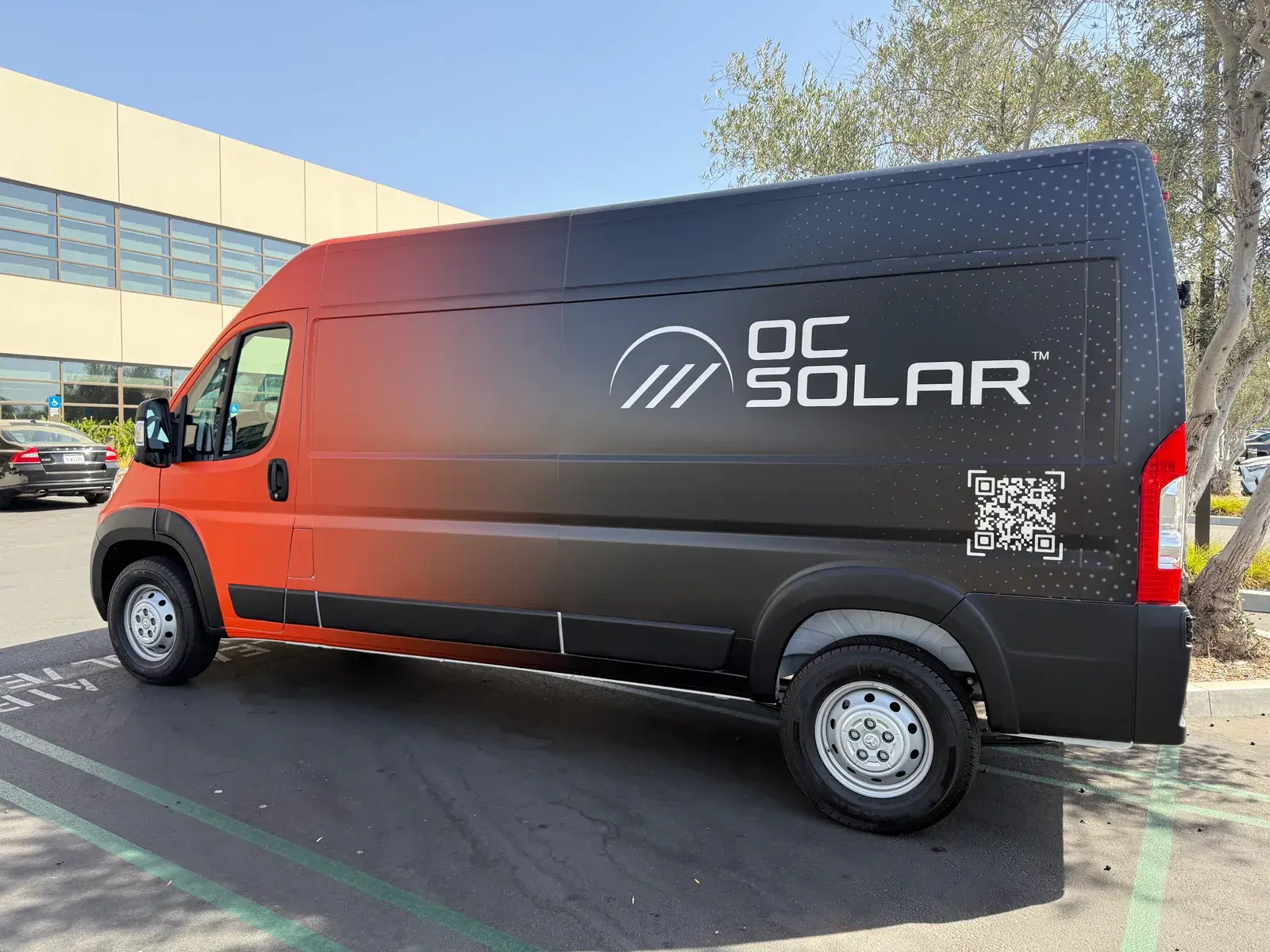 Side profile of a branded cargo van with an orange-to-black fade wrap, logo, and QR code parked outside an office building.