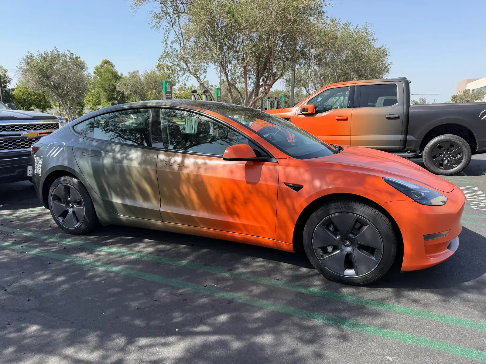 Side profile of a Tesla Model 3 sedan with an orange-to-gray fade wrap parked in a lot beside an orange pickup truck near EV chargers.