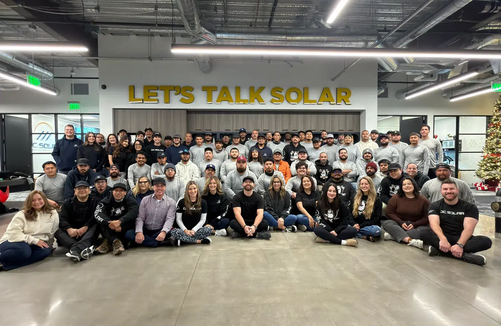 Dozens of OC Solar employees pose together, seated and standing, in front of a gold LET'S TALK SOLAR sign inside the office with a Christmas tree nearby.