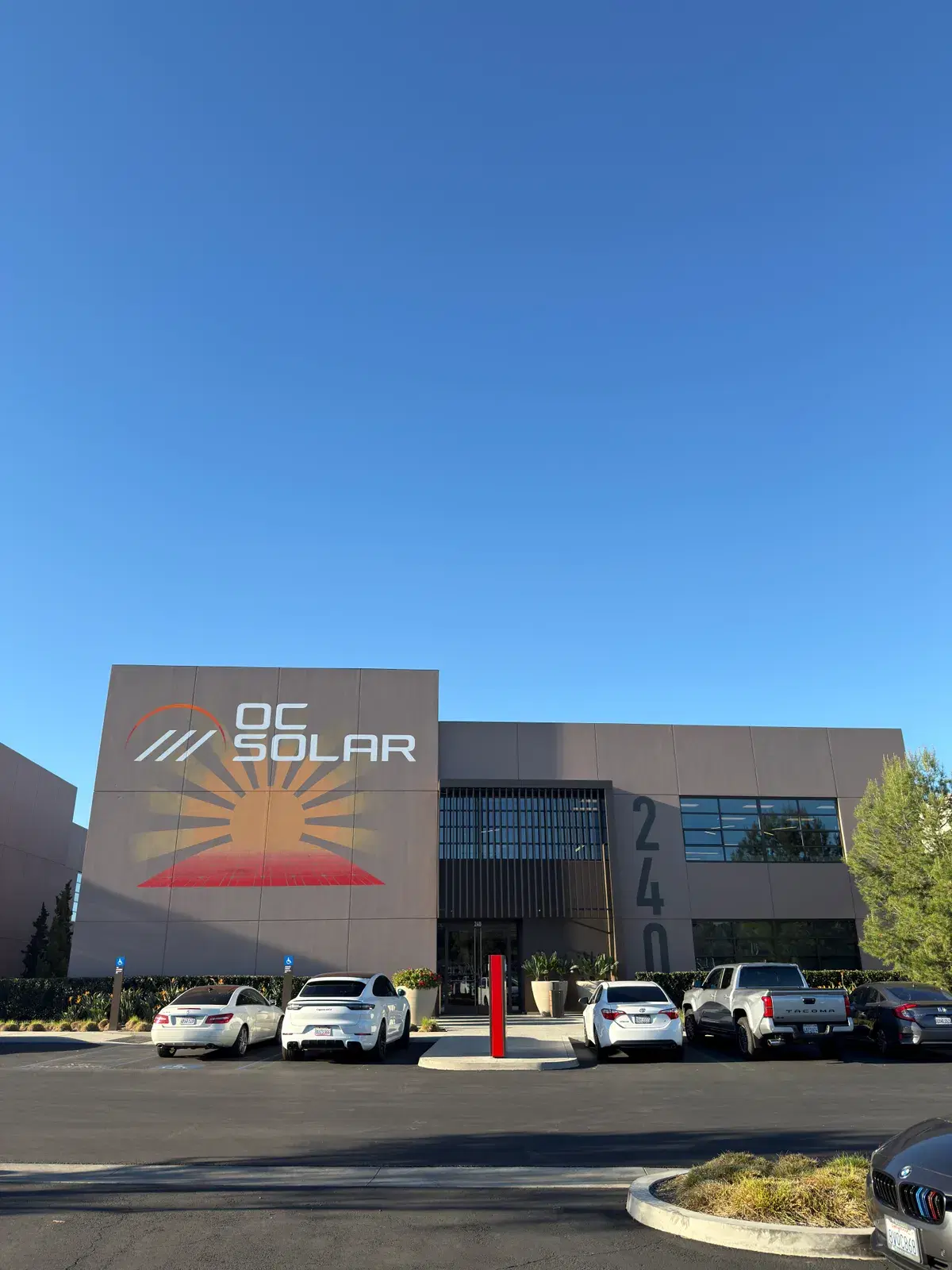 Exterior of the OC Solar office building showing the branded sunrise logo and parked cars out front.