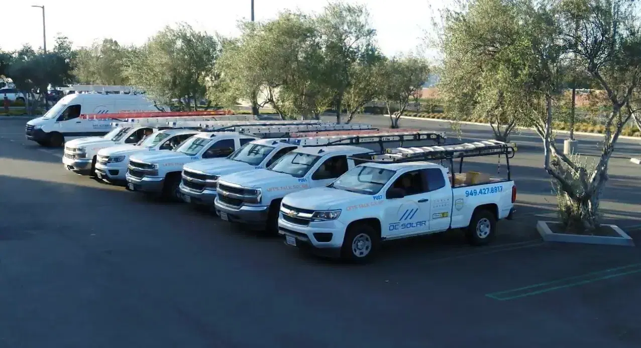 A row of white OC Solar branded Chevrolet pickup trucks with ladder racks and a van parked in a lot.