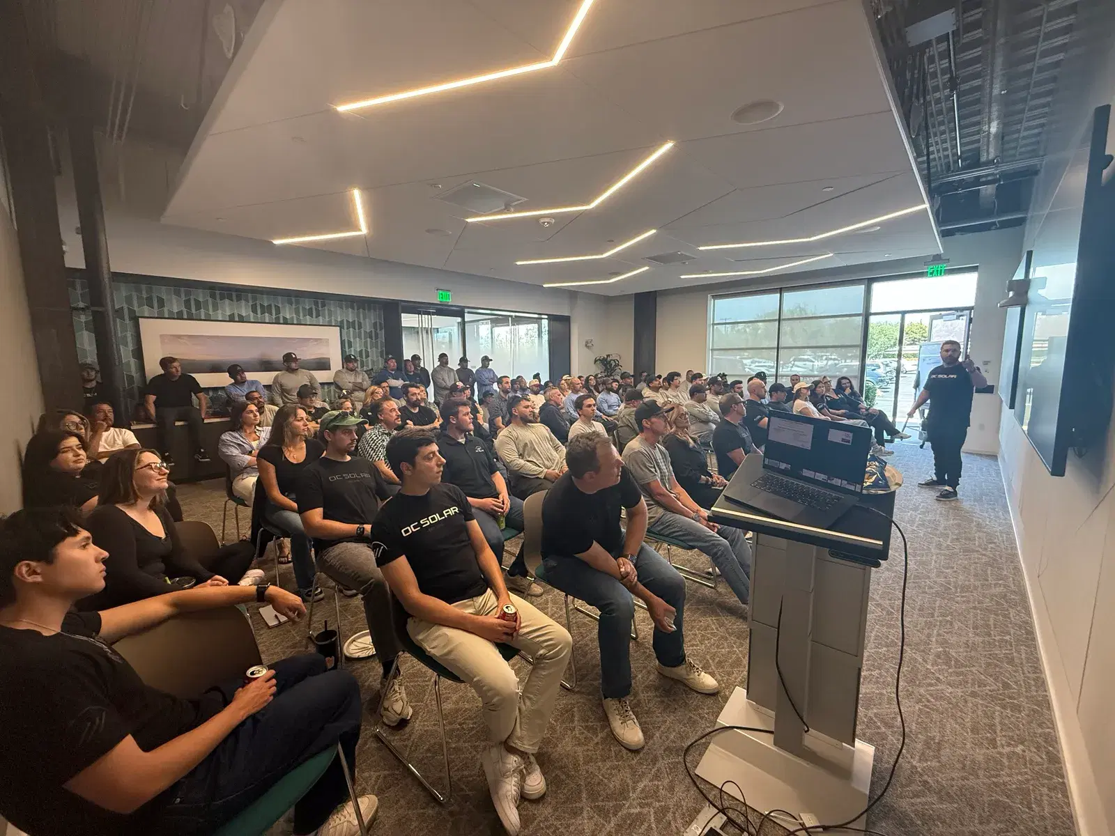 A large group of seated employees, several in branded OC Solar shirts, watching a presenter at a podium in a modern office room.