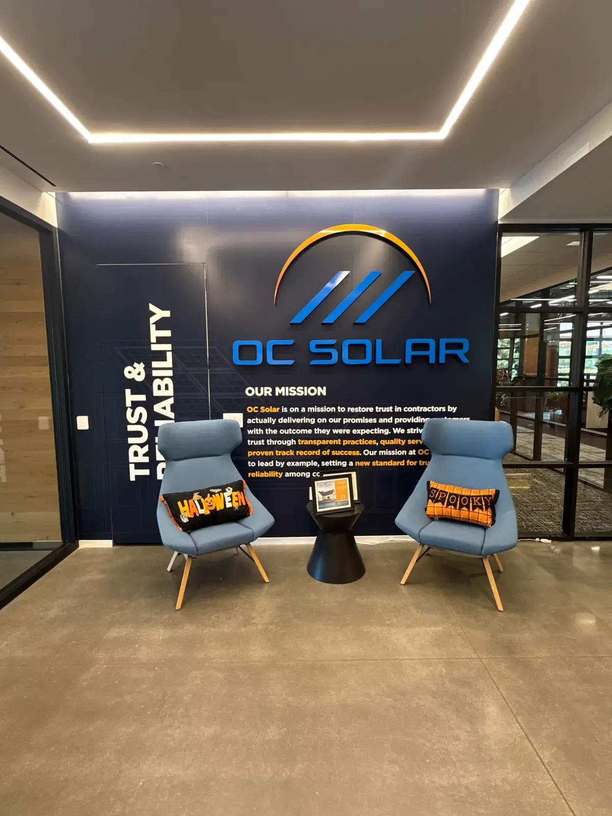 Office lobby featuring a dark wall with the OC Solar logo and company mission text, flanked by blue lounge chairs on a wood floor.