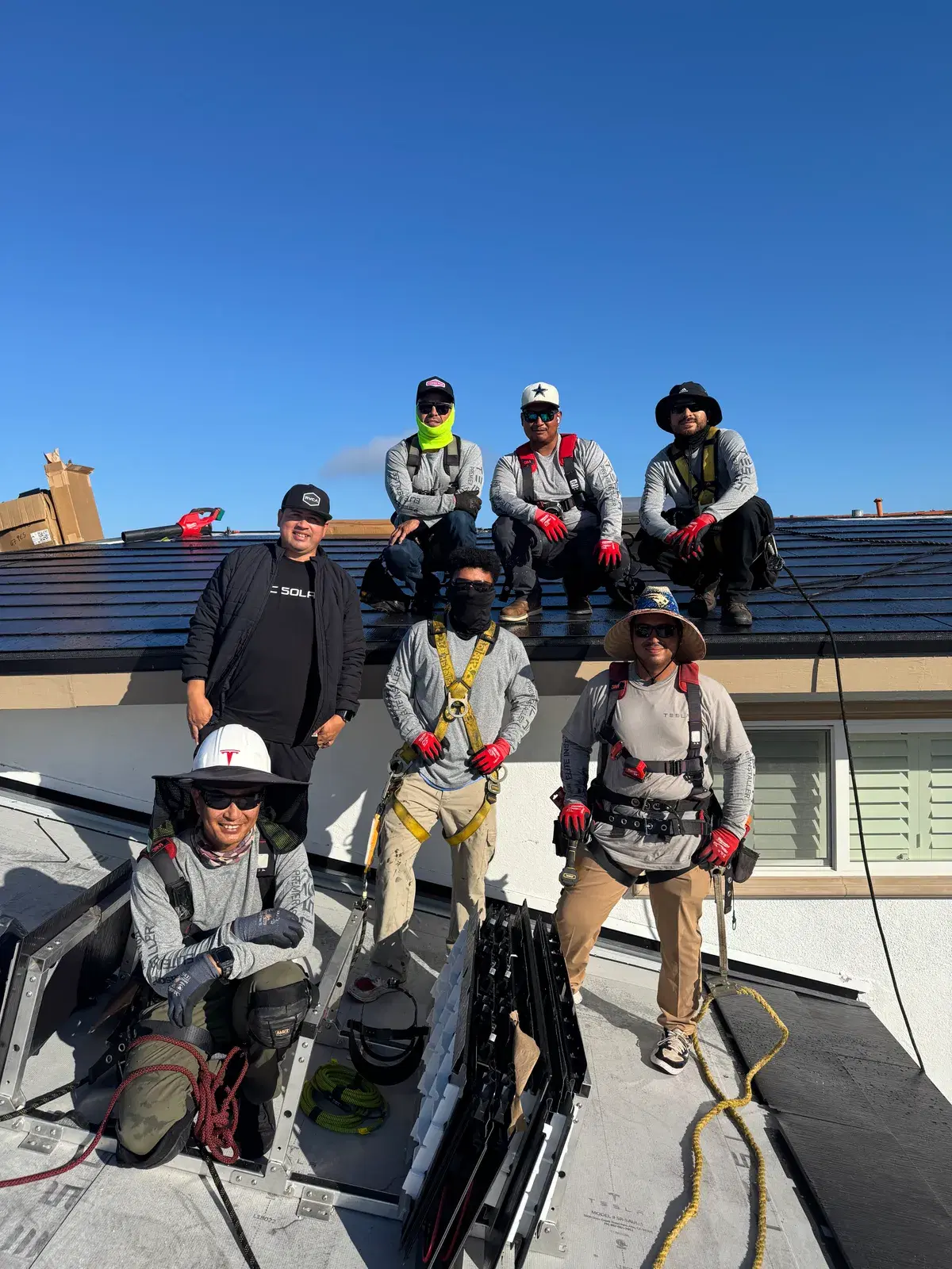 A team of solar installers in safety gear pose together on a rooftop under a clear blue sky, roofing materials nearby.