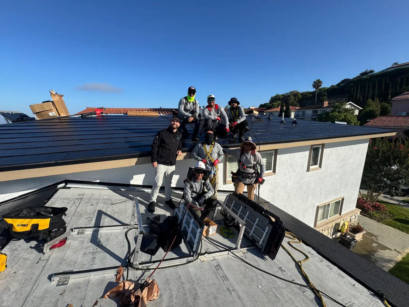 Seven OC Solar workers in safety harnesses pose on a roof beside a tile hoist, dark Solar Roof tiles and blue sky behind them.