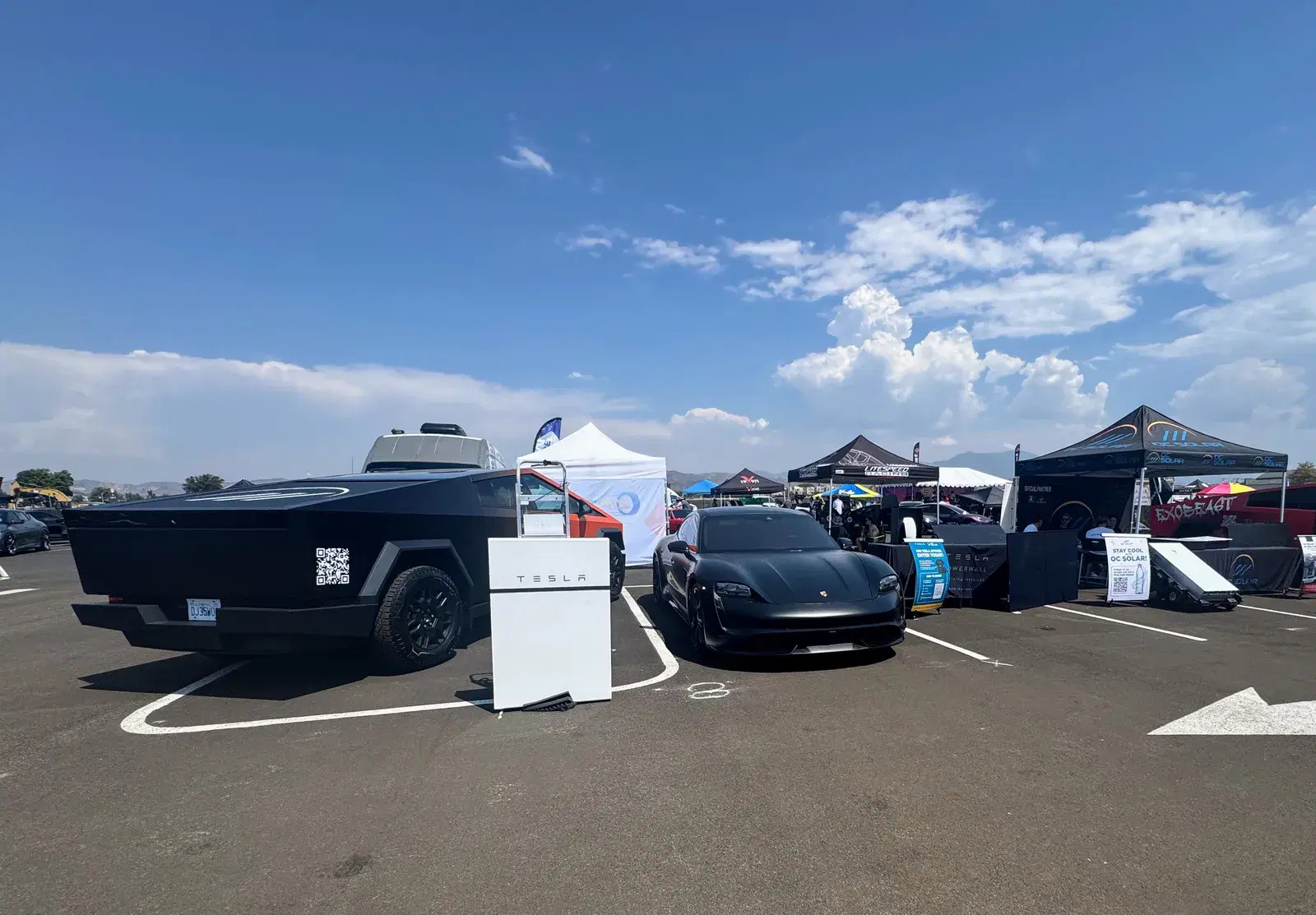 Outdoor event lot with a Tesla Cybertruck, a black sports car, branded tents, and a Tesla Powerwall on display under a blue sky.
