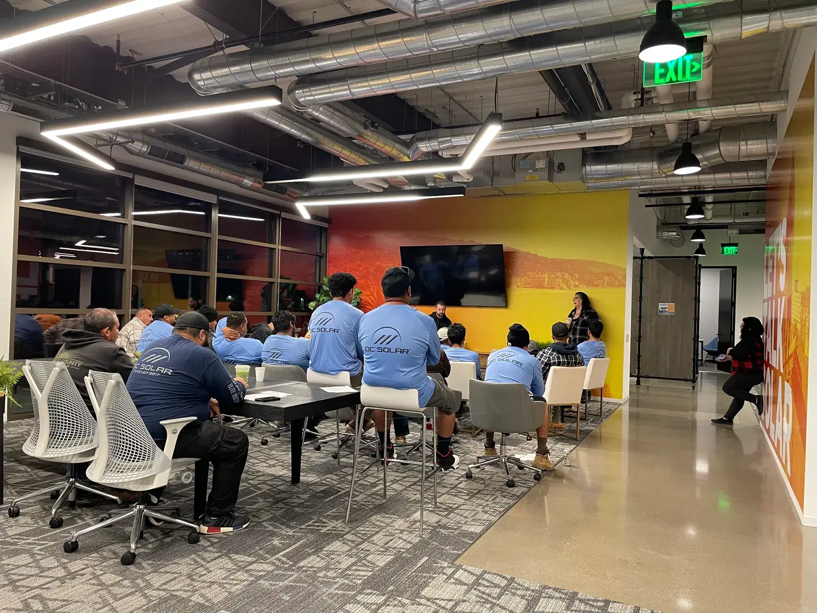 Employees in matching blue OC Solar shirts seated around a table during a meeting in a modern open office.