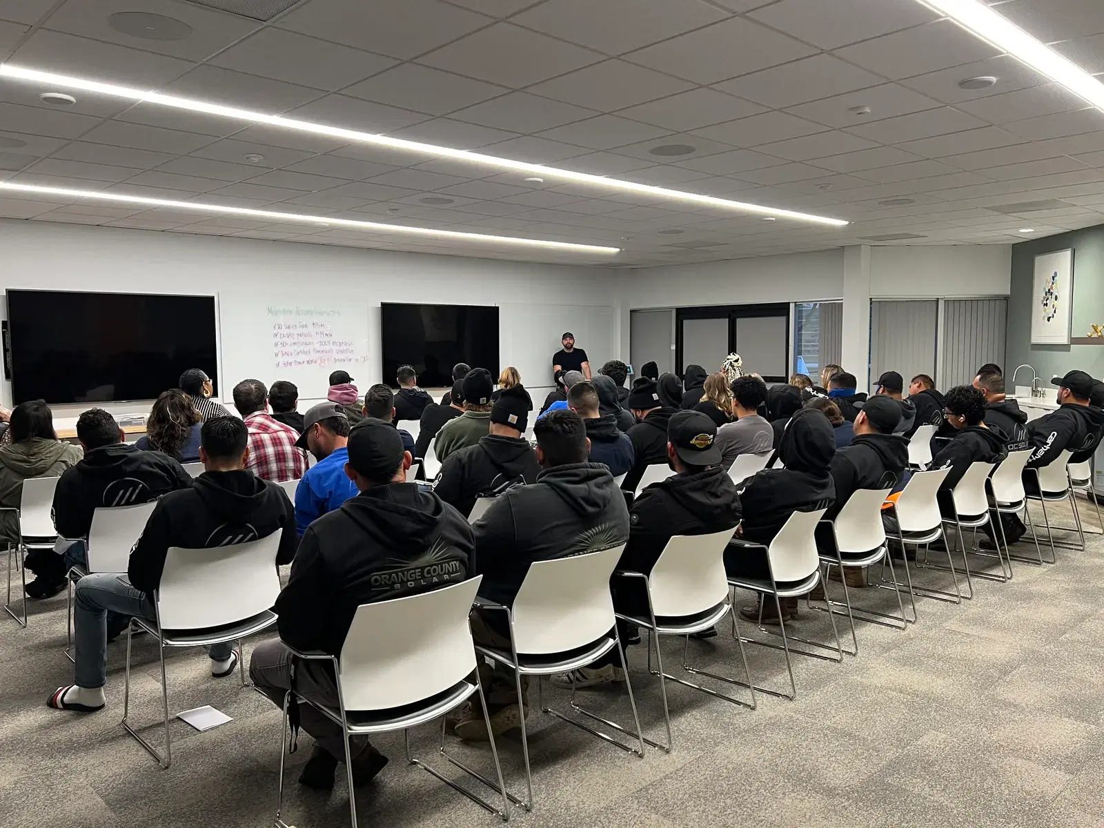 A large group of employees in dark Orange County Solar hoodies seated in rows facing a presenter in a training room.