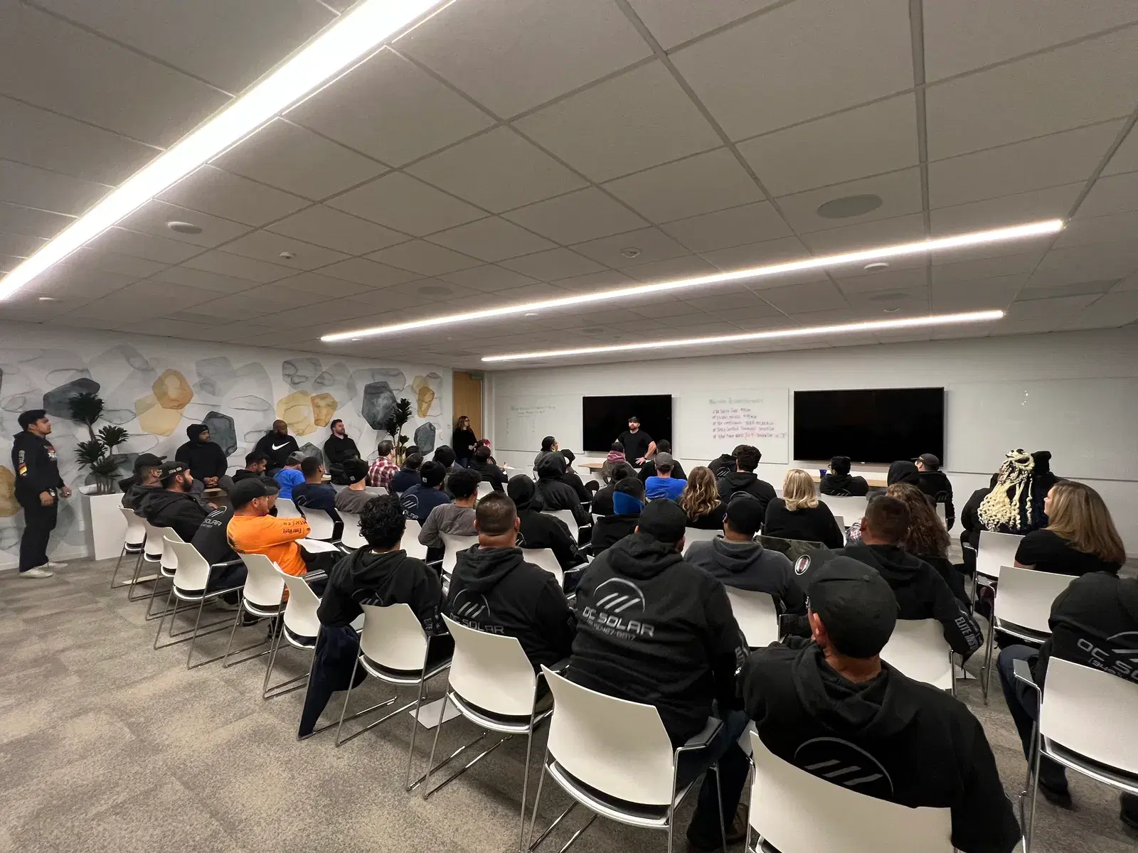 Employees wearing OC Solar branded hoodies seated in a bright training room with a decorative stone wall, listening to a presenter.