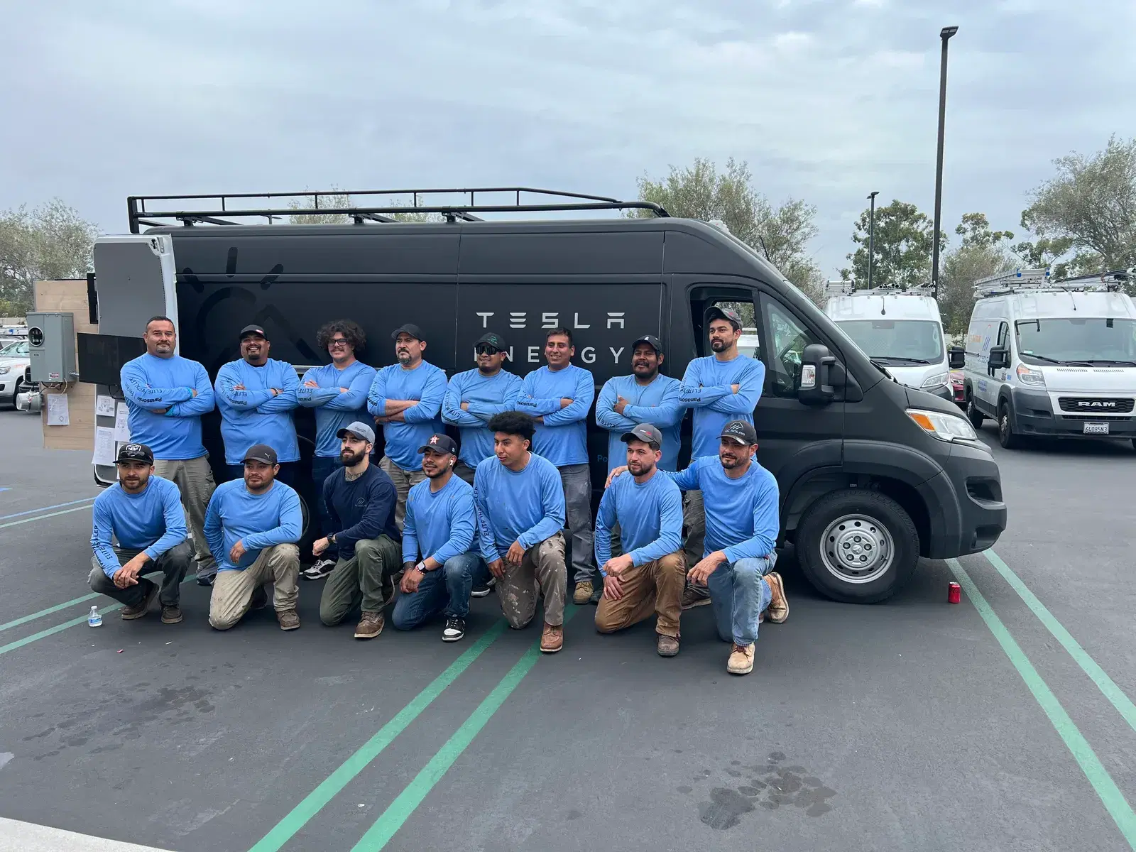 A group of solar installers in blue shirts posing in front of a black Tesla Energy work van in a parking lot.