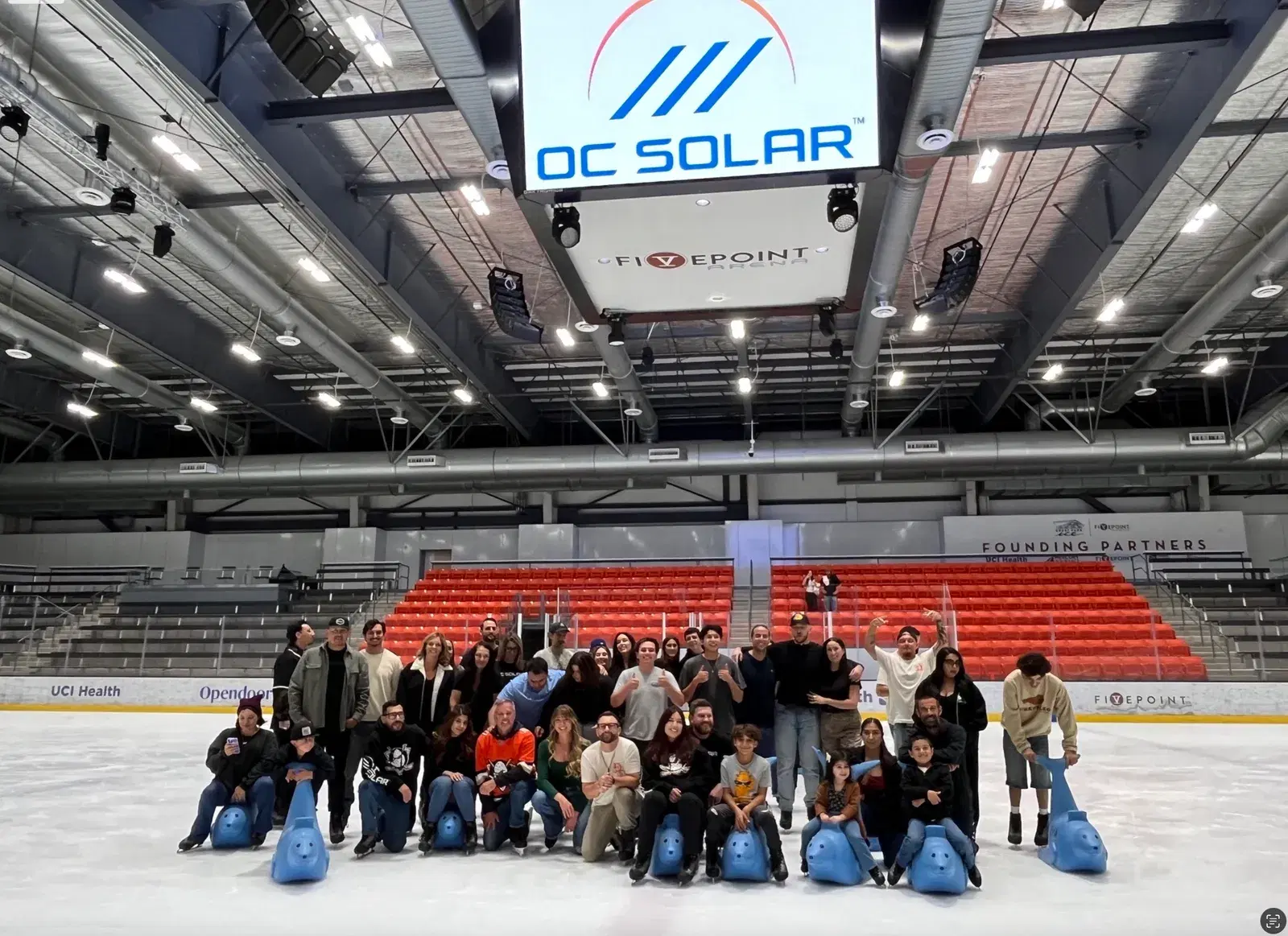 Large group of OC Solar employees posing on an indoor ice rink under a jumbotron displaying the OC Solar logo.