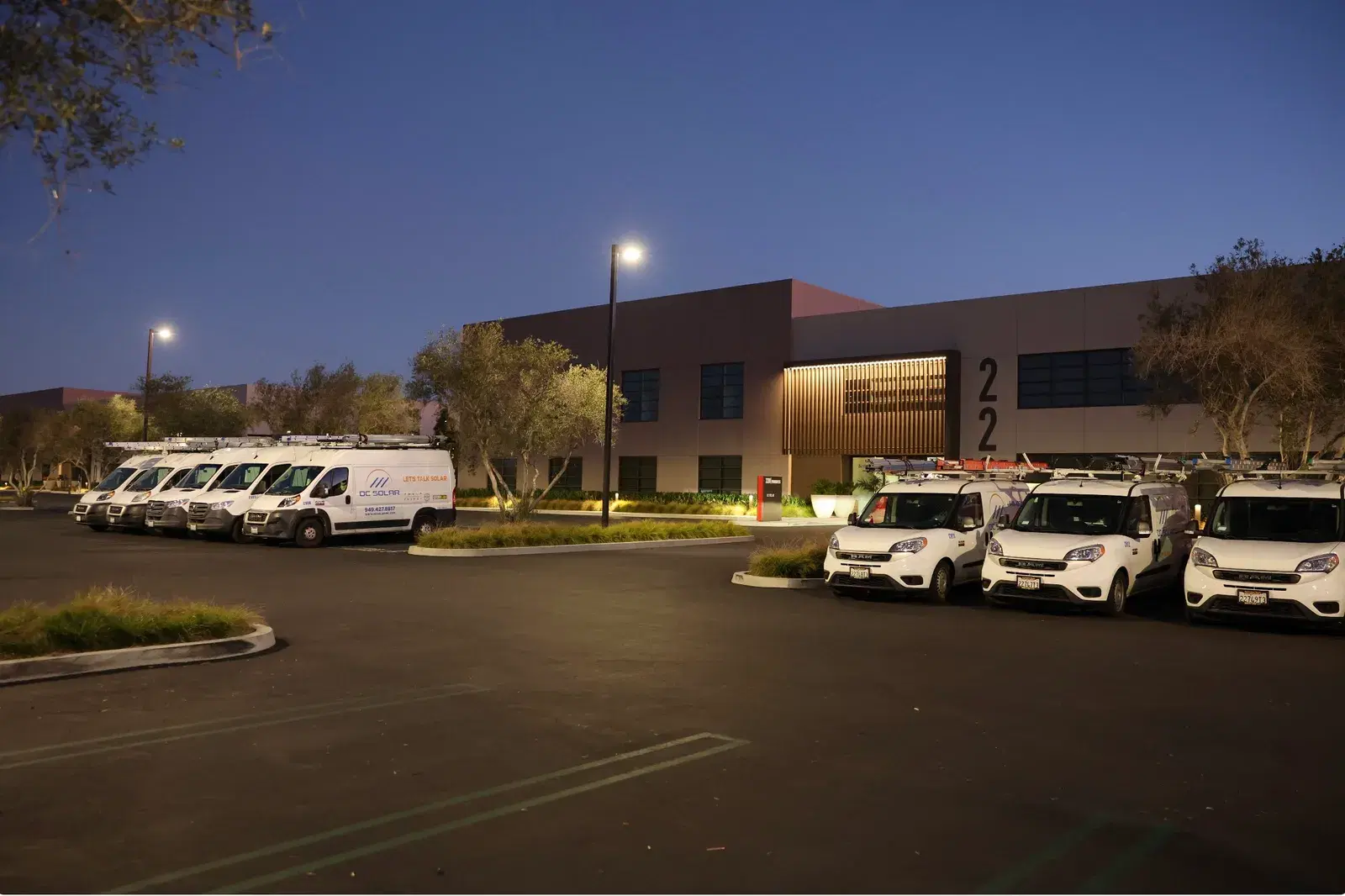 Rows of white OC Solar branded vans and cargo vans parked outside the company office building at dusk.