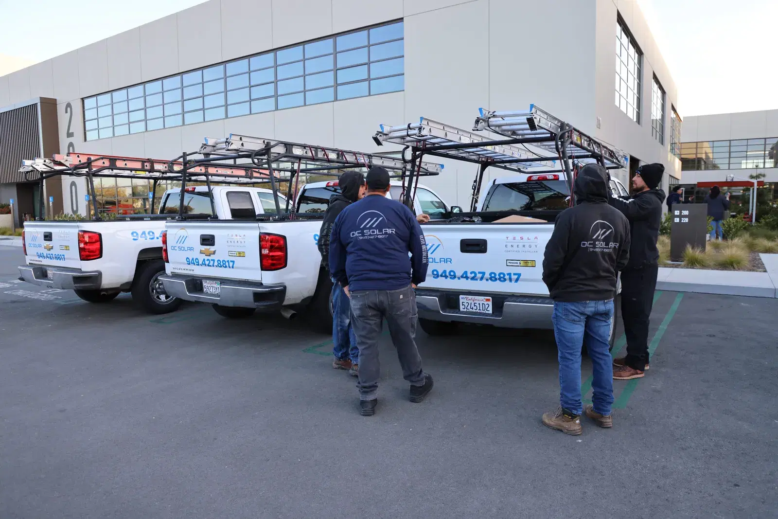 Installers standing beside a fleet of white OC Solar pickup trucks with ladder racks parked outside the company building.