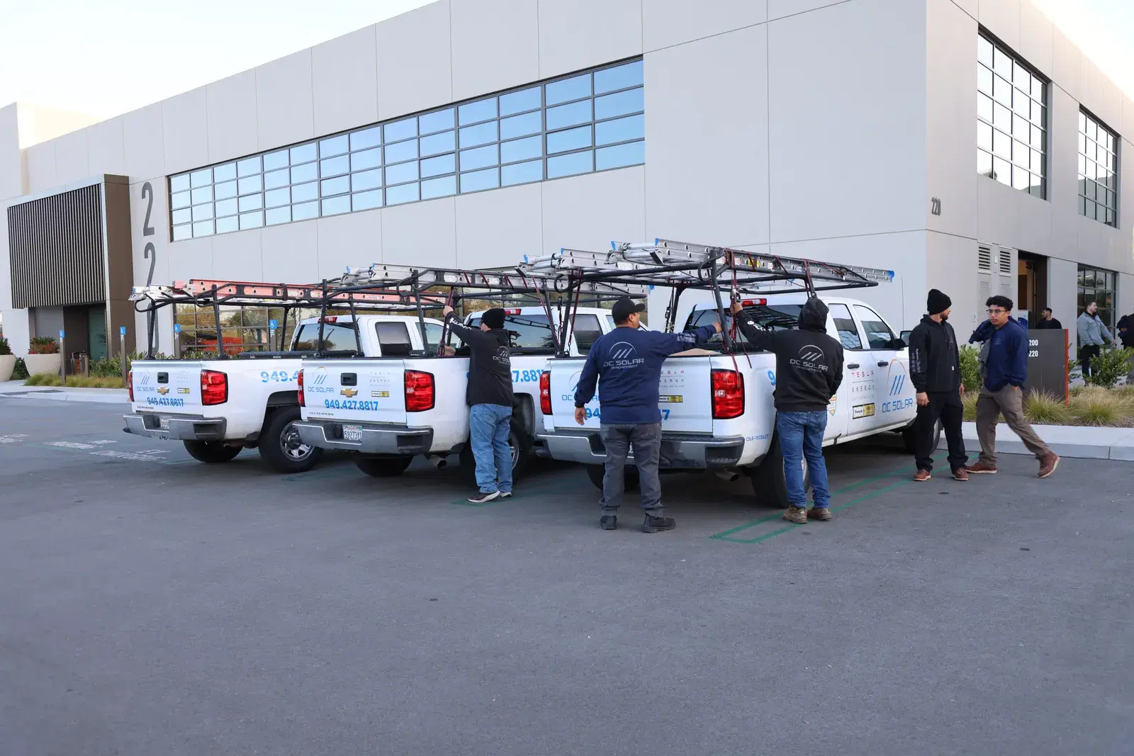 A line of branded white OC Solar pickup trucks with ladder racks parked outside the company building as workers prepare to load equipment.