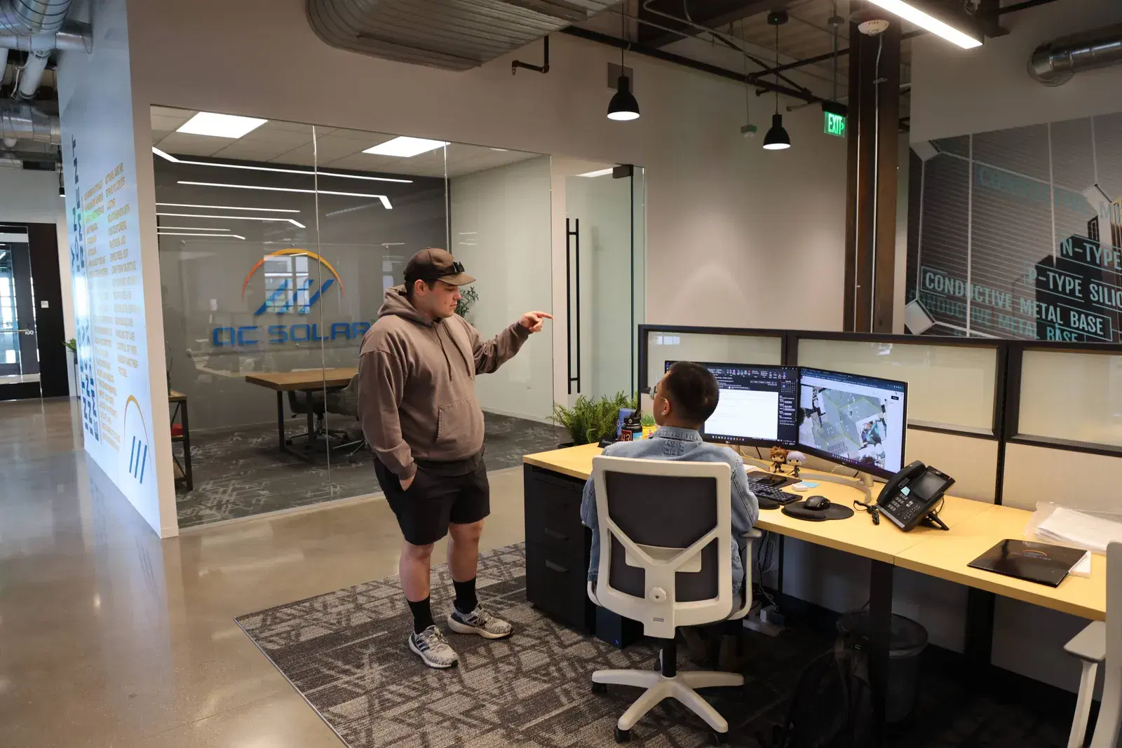 Two OC Solar employees collaborating at a desk with dual monitors inside a modern office, the company logo on a glass wall behind them.