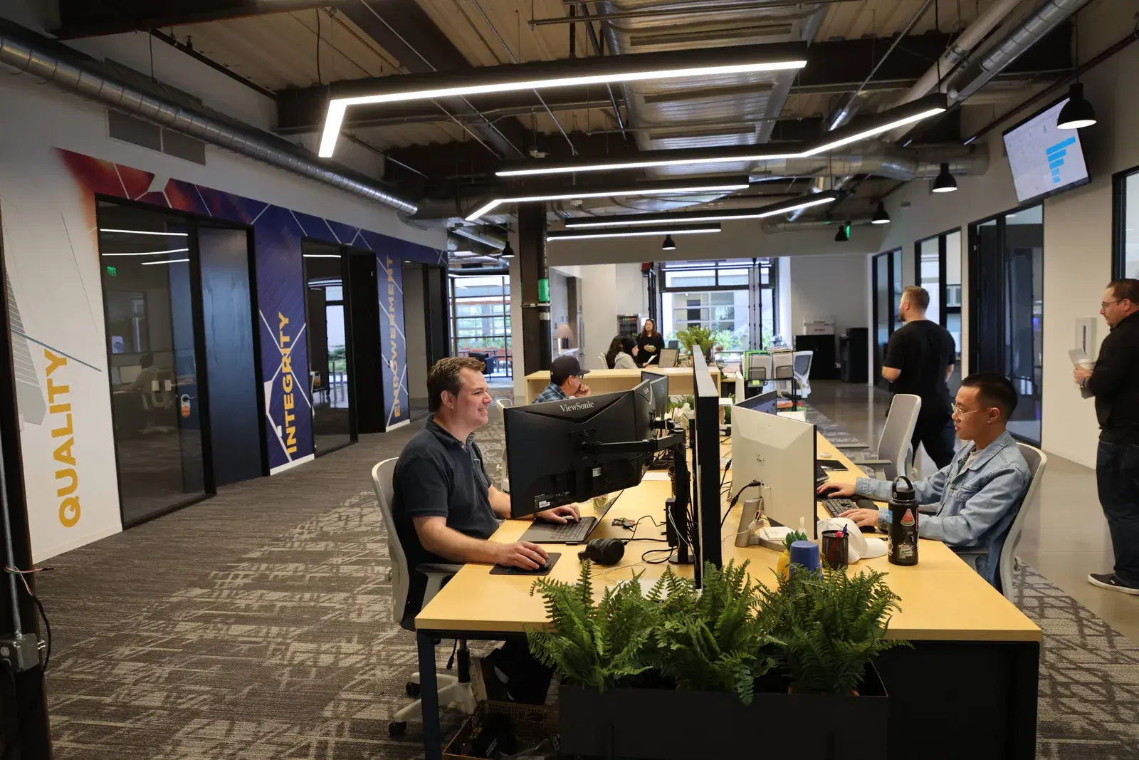 Employees working at computers in a modern open-plan office with Quality and Integrity printed on the walls.