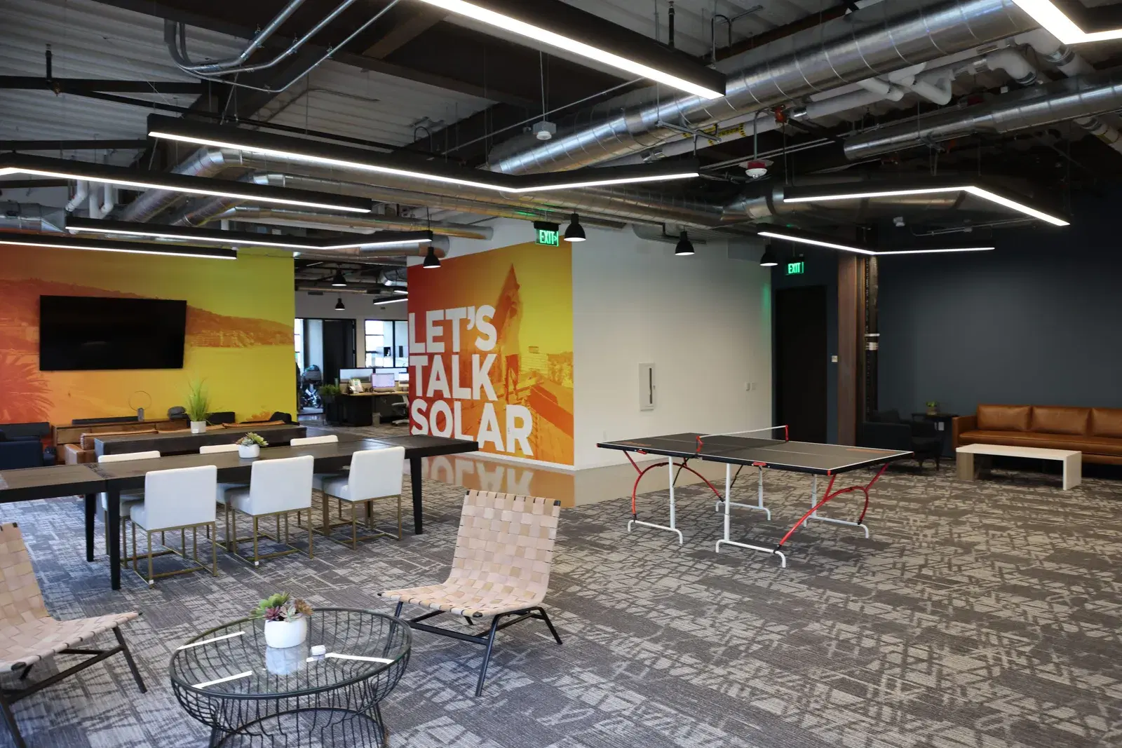 Bright office lounge with exposed-ceiling ductwork, a yellow-and-orange solar-themed wall mural, dining tables, a ping-pong table, and woven lounge chairs.