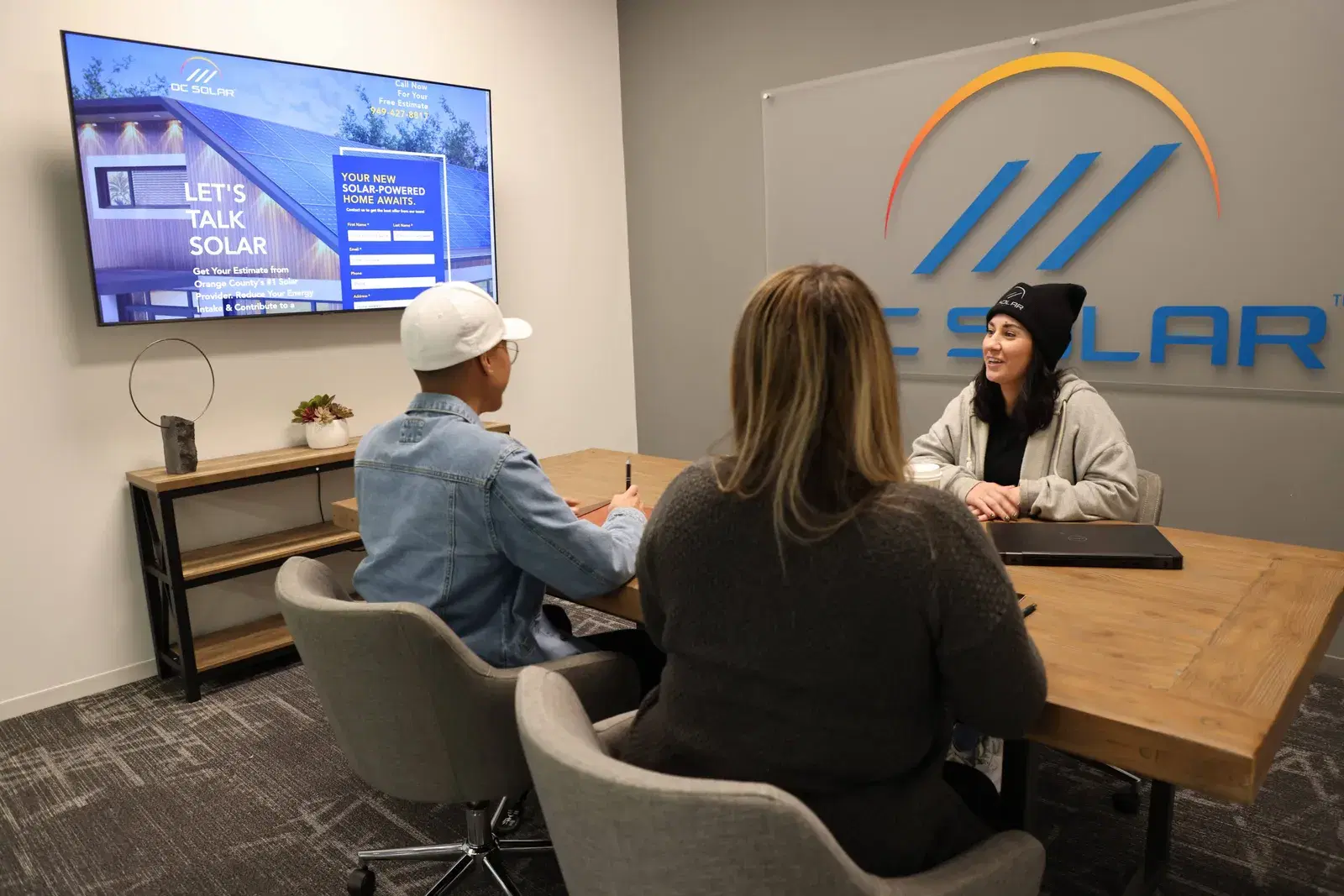Three people seated at a wooden table in a meeting room, a wall-mounted screen showing a solar ad and a large OC Solar logo on the wall behind them.