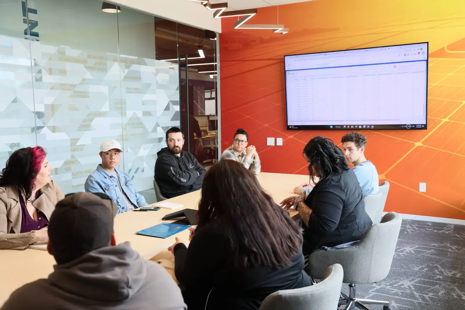 A group of employees seated around a long conference table looking at a wall-mounted monitor displaying a spreadsheet, with an orange solar-graphic feature wall.