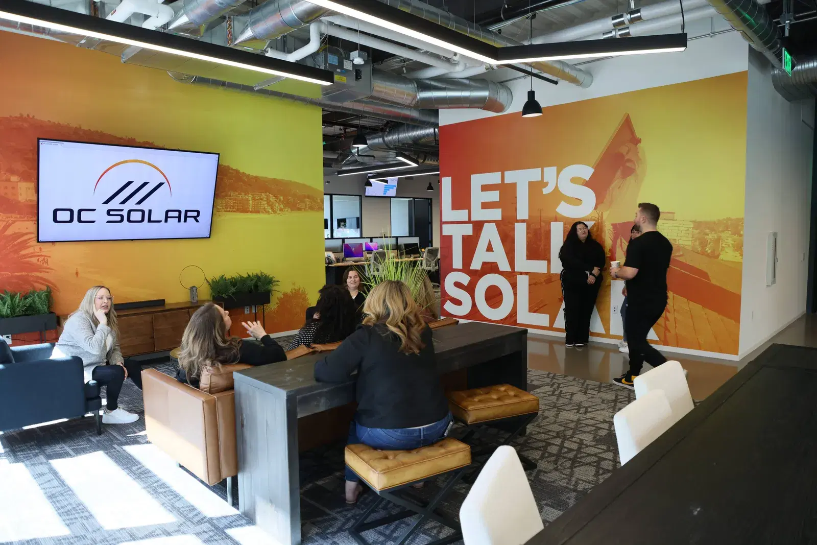 Several employees sitting and standing in an open office lounge, with an OC Solar logo on a wall screen and a large orange mural reading Let's Talk Solar.