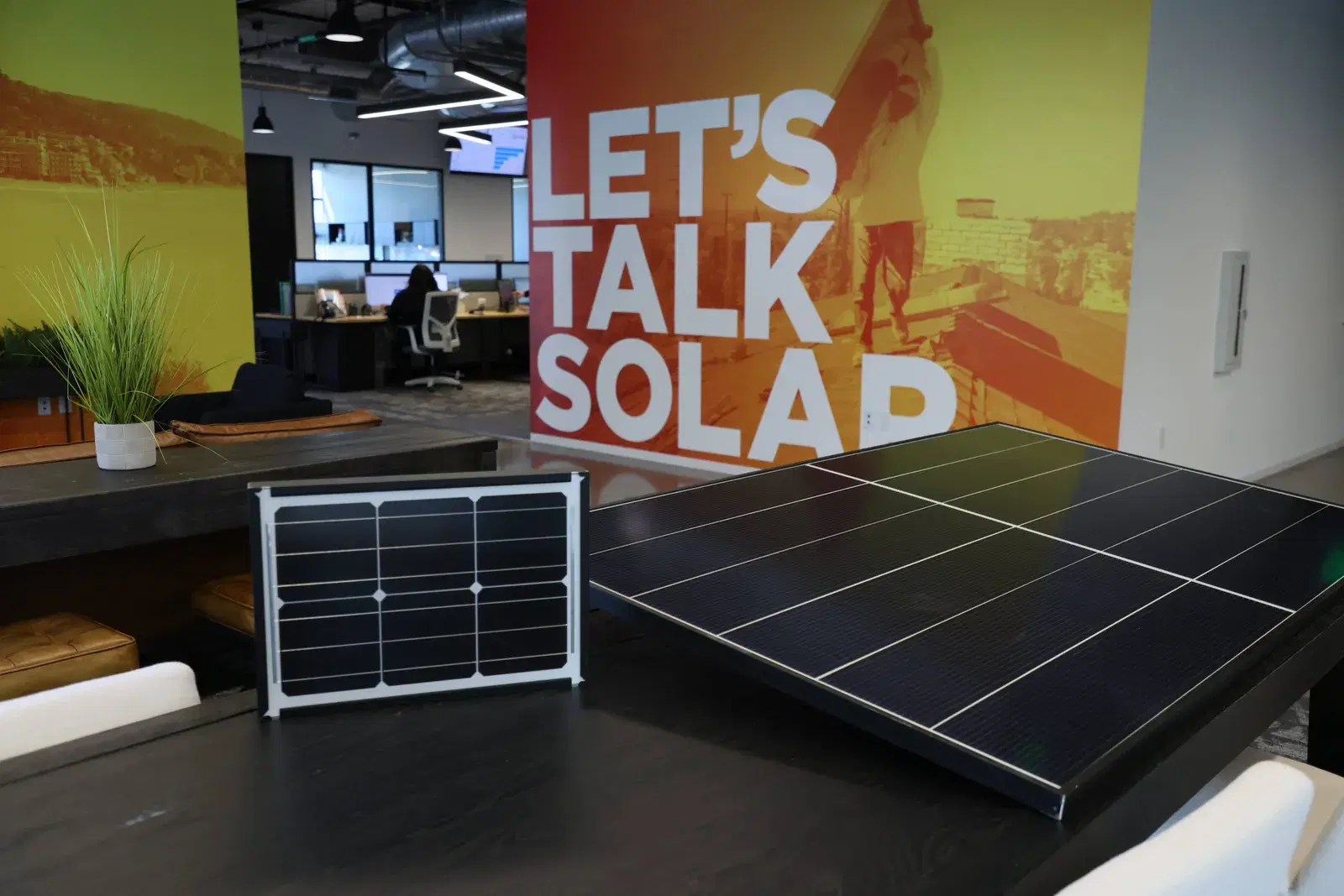 A large black solar panel and a small framed demonstration solar panel on a dark table in the OC Solar office, with a Let's Talk Solar mural and a team member at a desk in the background.