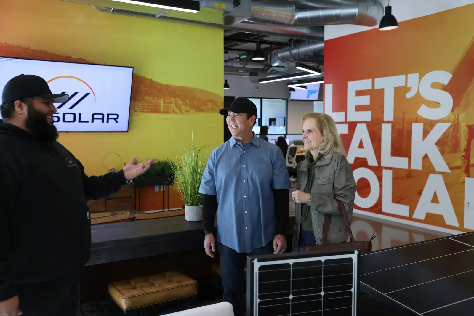 A smiling employee gesturing toward a man and woman in an office showroom, with an OC Solar logo screen, a Let's Talk Solar mural, and a demo solar panel nearby.