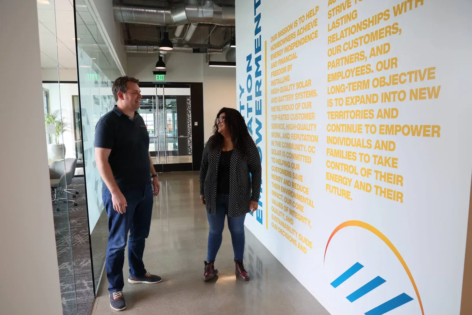 A man and a woman talking and smiling in an office corridor next to a wall printed with the company mission statement and the OC Solar logo graphic.