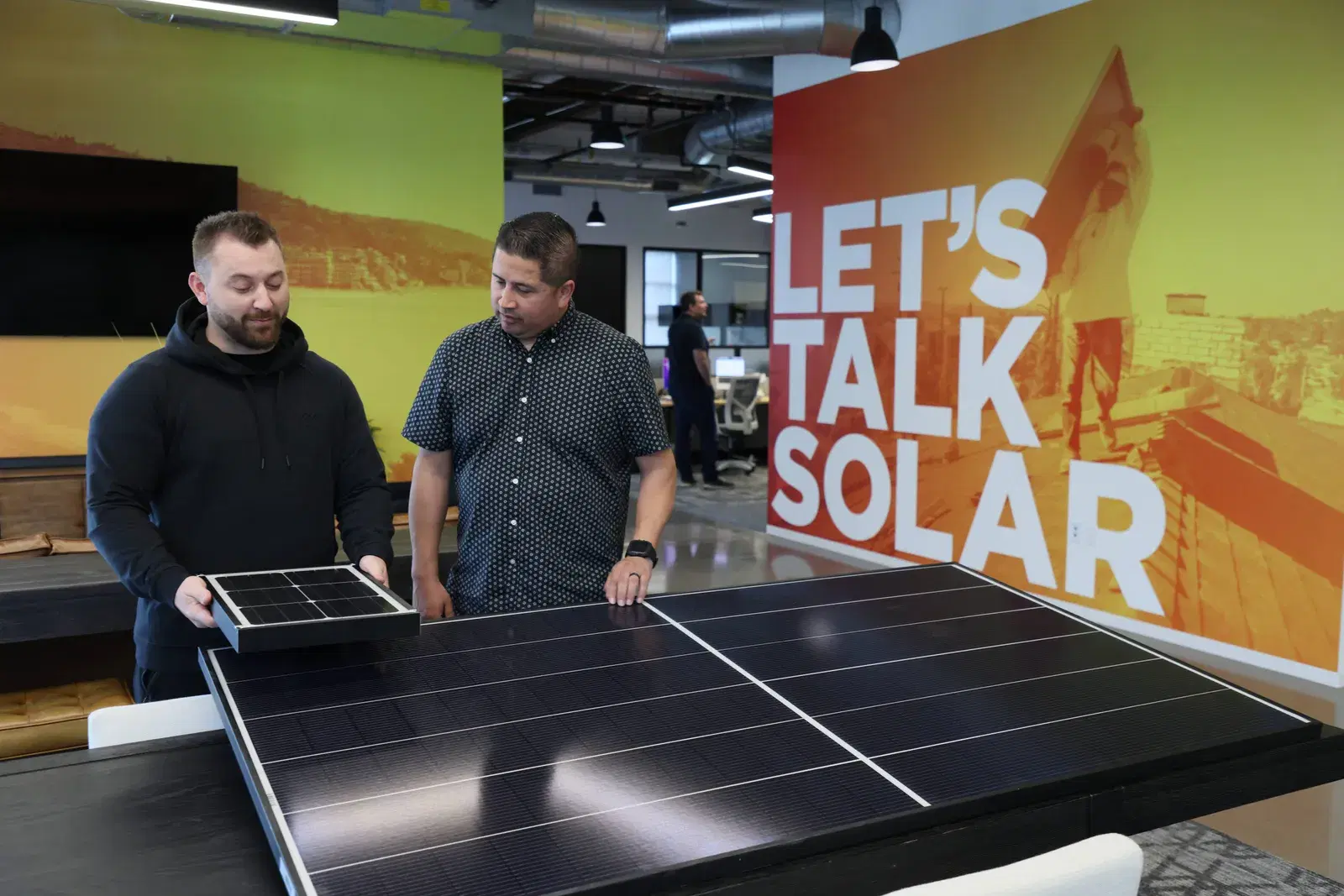 Two men examining solar panels in an office showroom, one holding a small demo panel beside a large black module, with a Let's Talk Solar mural behind them.