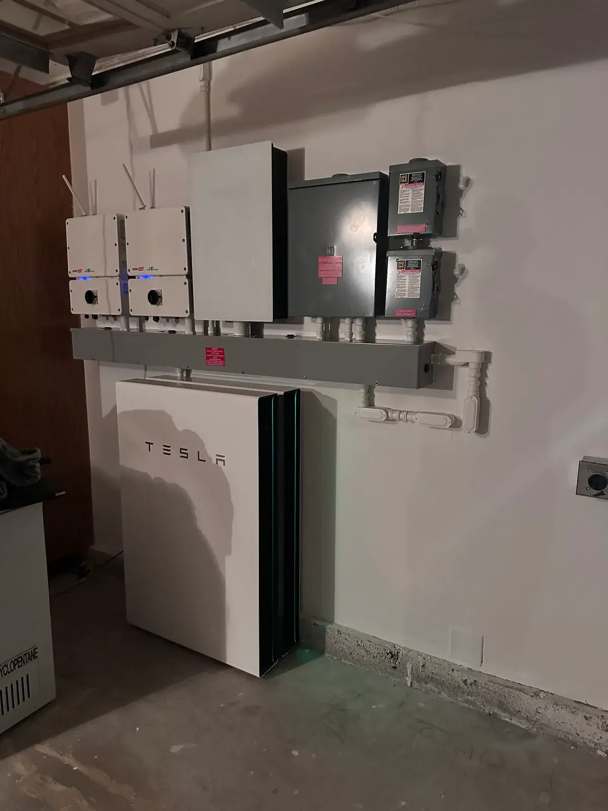 A white Tesla Powerwall battery mounted on a garage wall beneath a row of inverters, a gray gateway enclosure, and electrical disconnect boxes with conduit running between them.
